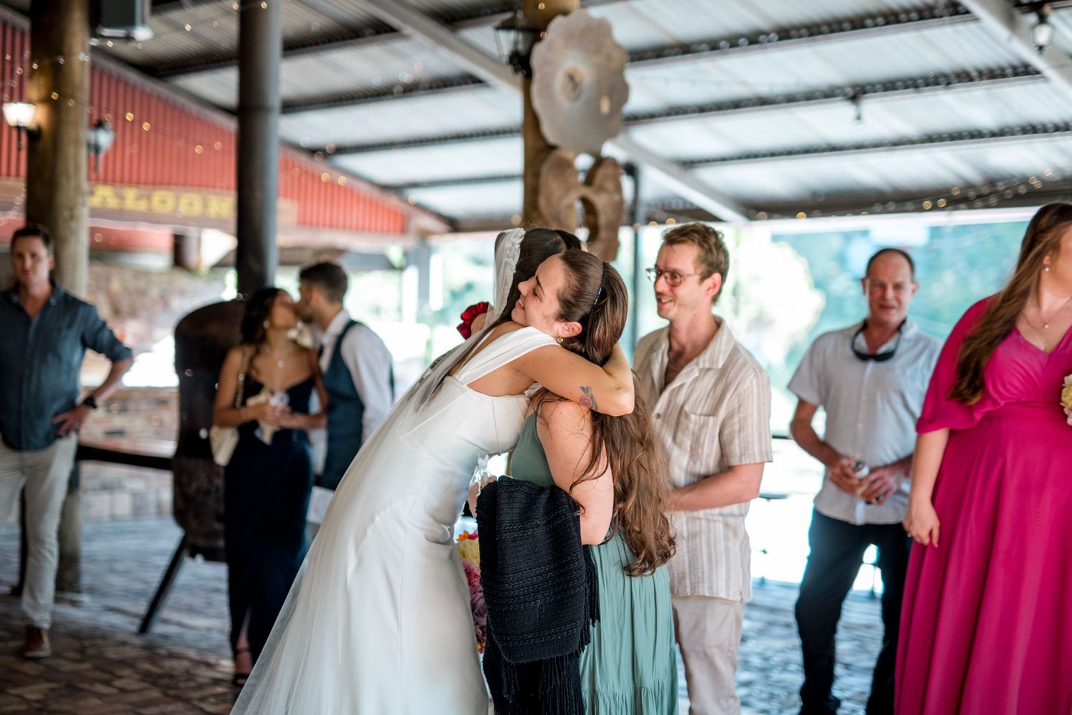 Bride in white gown shares warm embrace with guest at lively outdoor wedding reception venue.