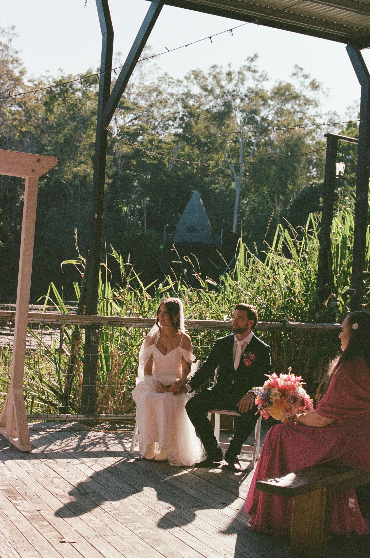 Bride and groom seated at outdoor wedding ceremony on wooden deck surrounded by greenery.