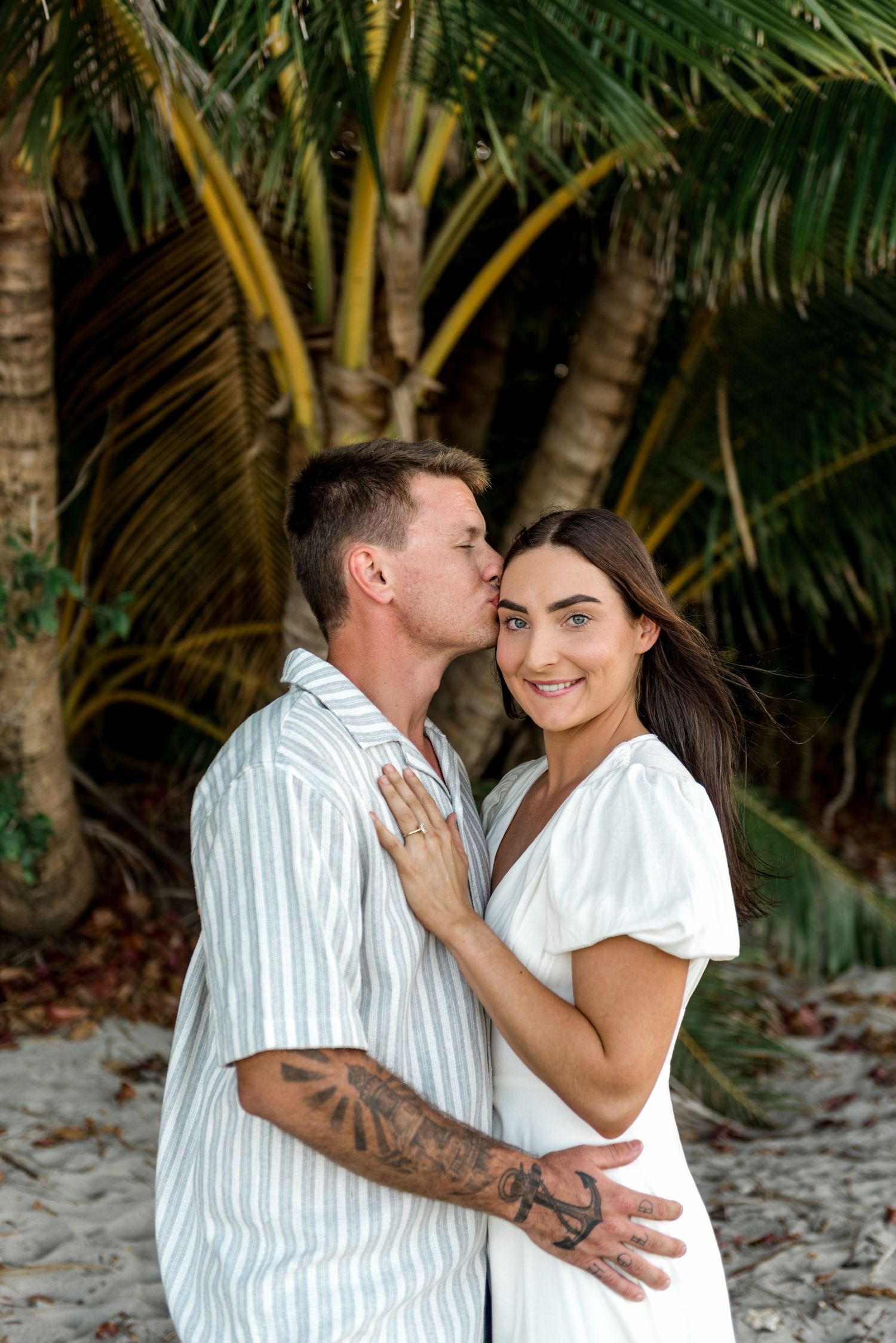 Man kissing womans cheek as she smiles at camera on tropical palm tree beach.