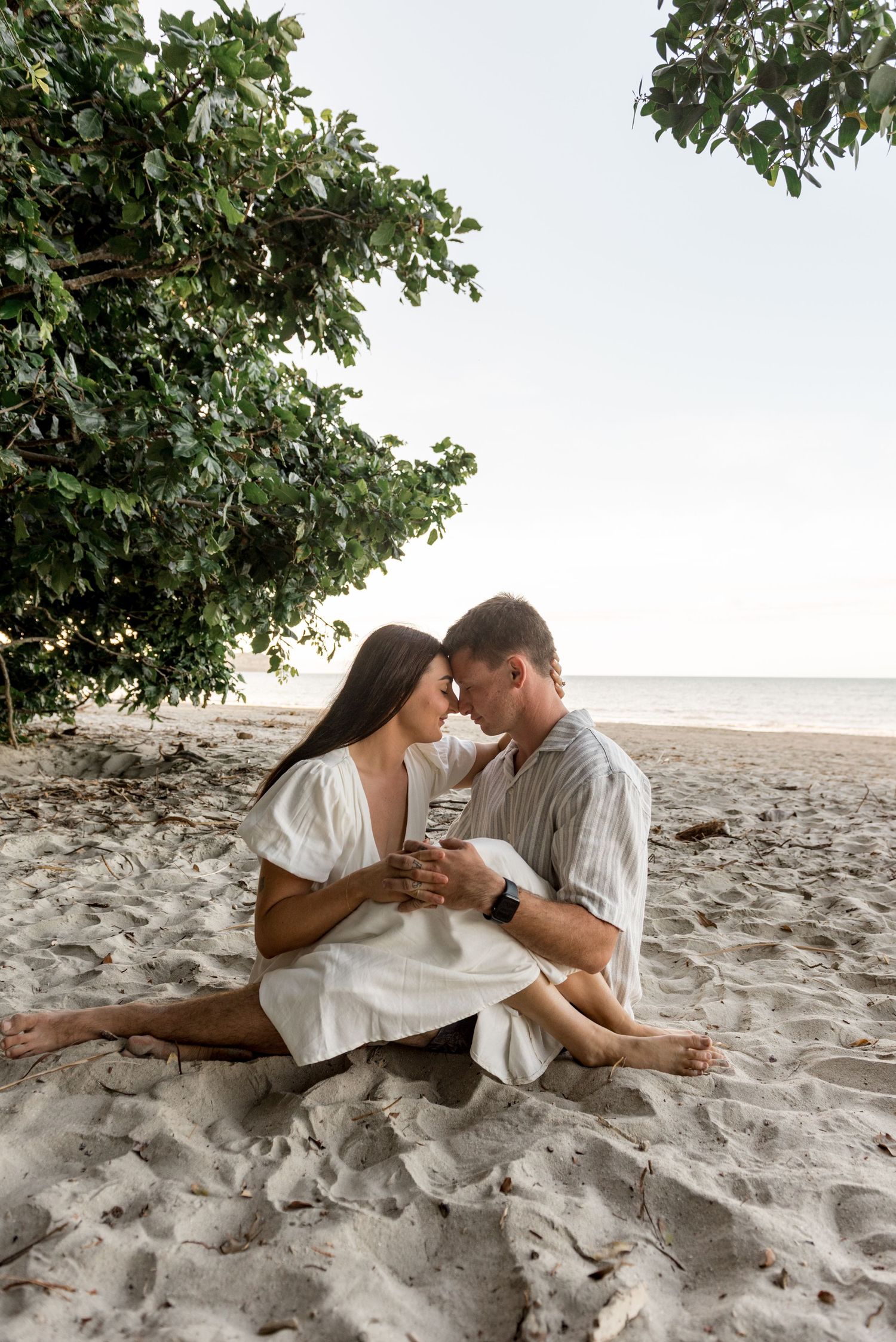 Couple sitting close together on sandy beach near the ocean, sharing an intimate moment under a green tree.