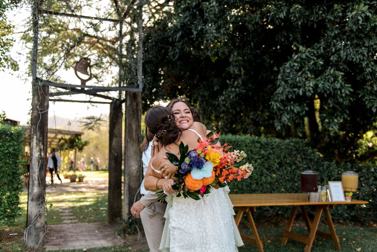 Bride holding colorful bouquet receives joyful hug from guest at outdoor wedding venue.
