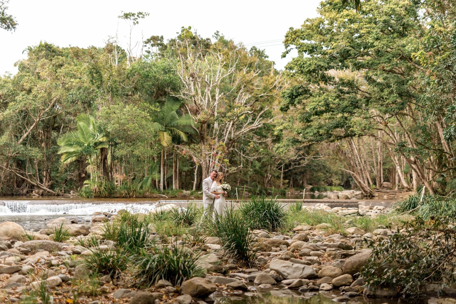 Bride and groom stand together among lush tropical greenery beside a rocky creek landscape.