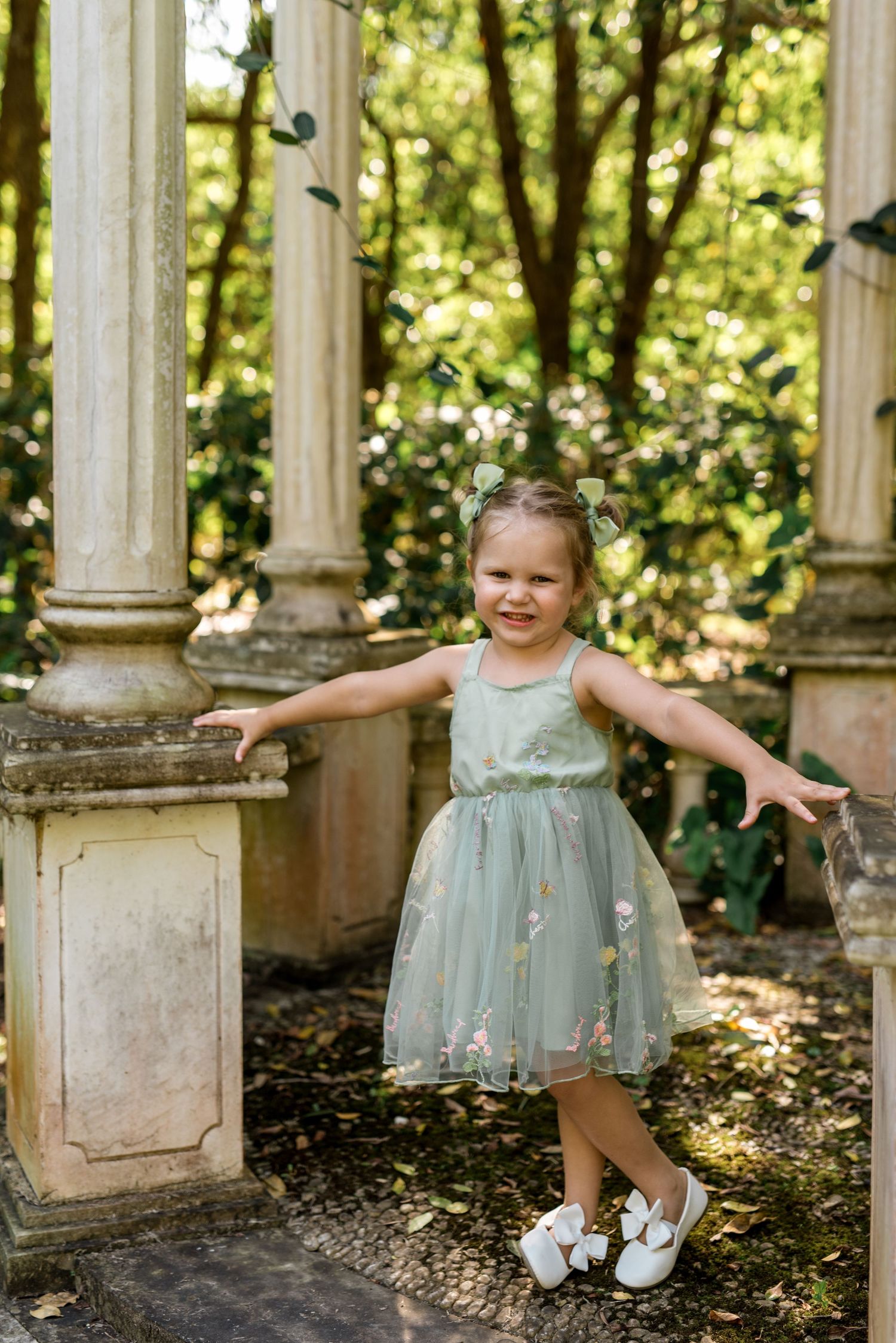 Young flower girl in mint green tulle dress posing playfully beside stone columns in garden setting.