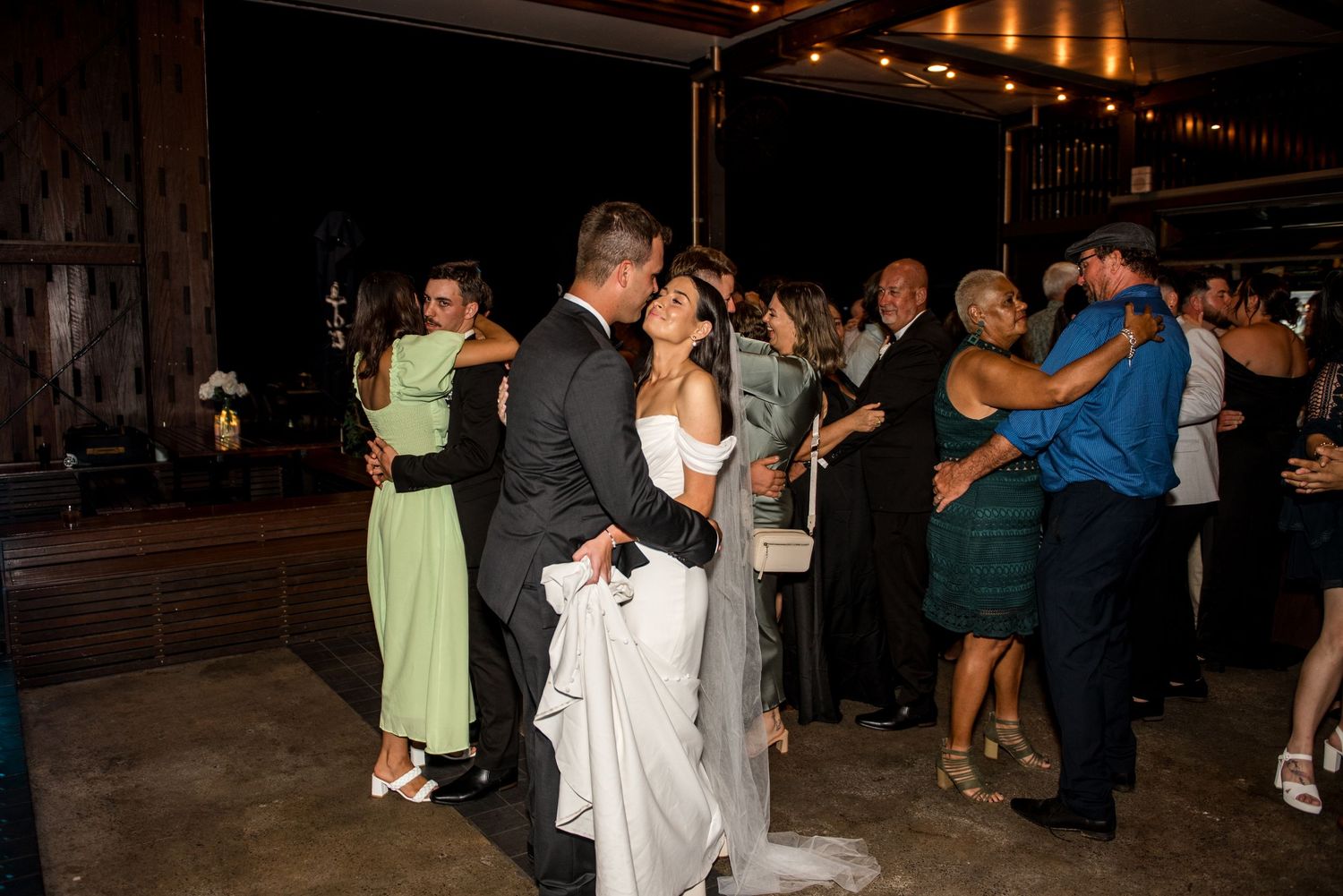 Bride and groom dance together surrounded by wedding guests on a dimly lit reception dance floor.