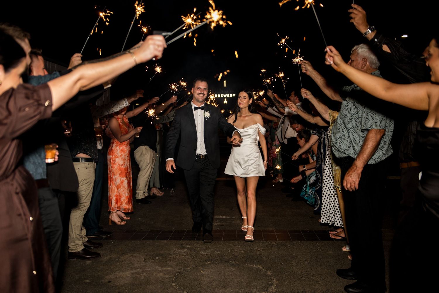 Bride and groom walk through sparkler tunnel surrounded by wedding guests at night.