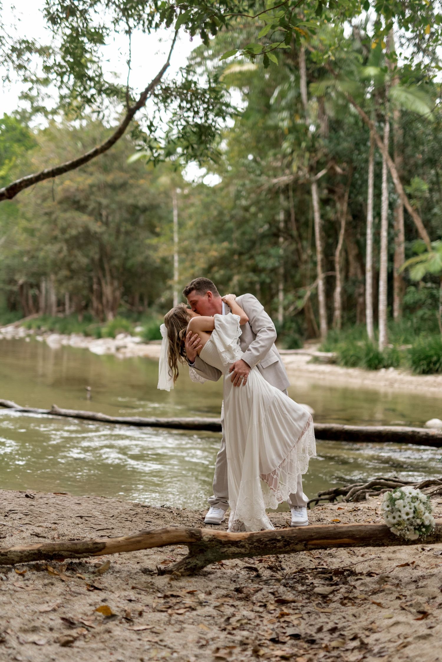 Groom dips bride for romantic kiss beside a forest river after outdoor wedding ceremony.