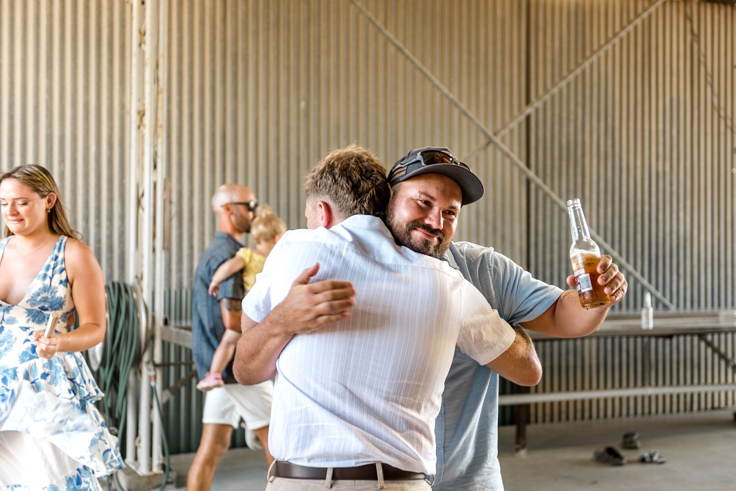Guest in cap hugs friend while holding beer at casual outdoor barn wedding.