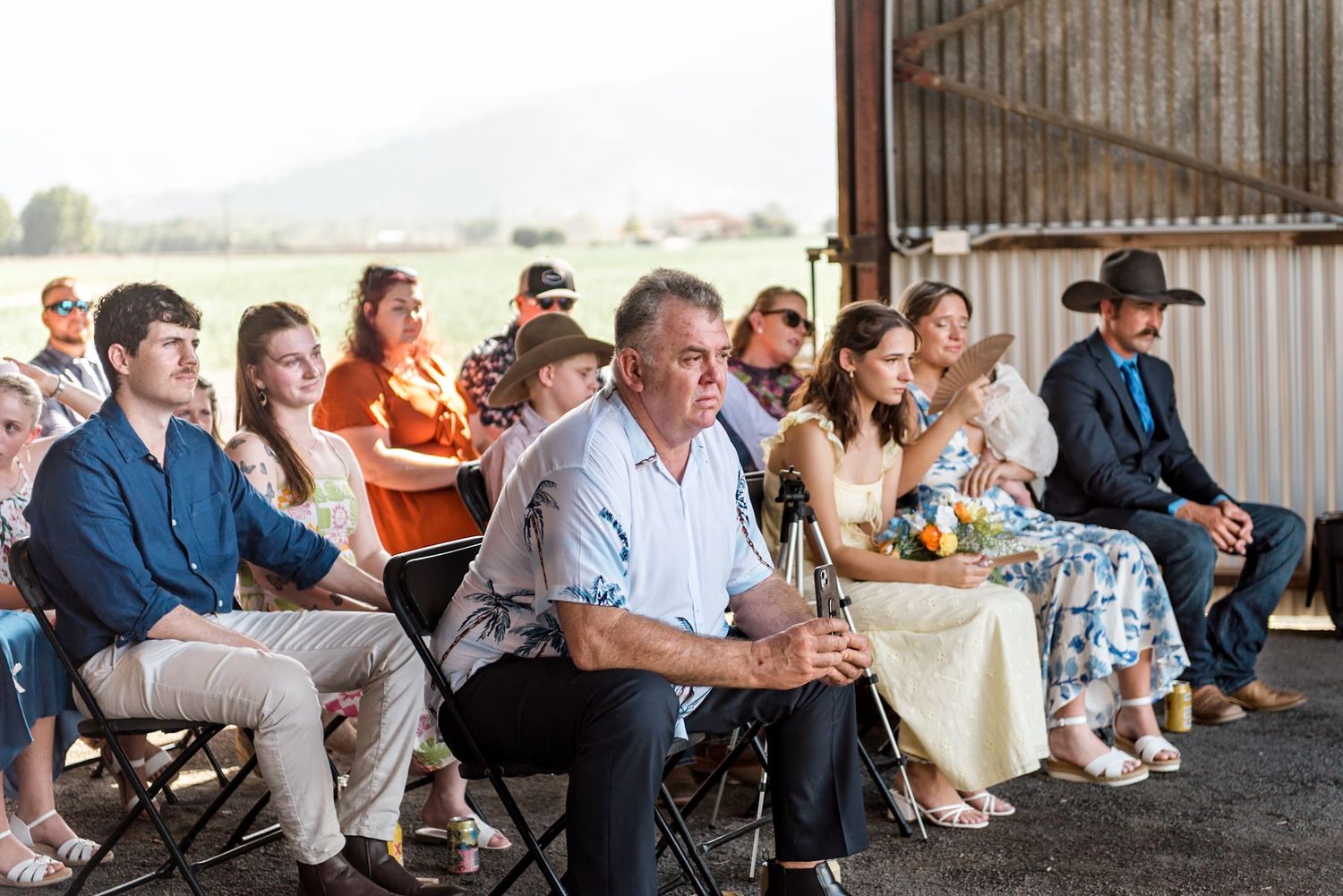 Diverse wedding guests seated in chairs watching ceremony inside rustic barn with green countryside visible outside.