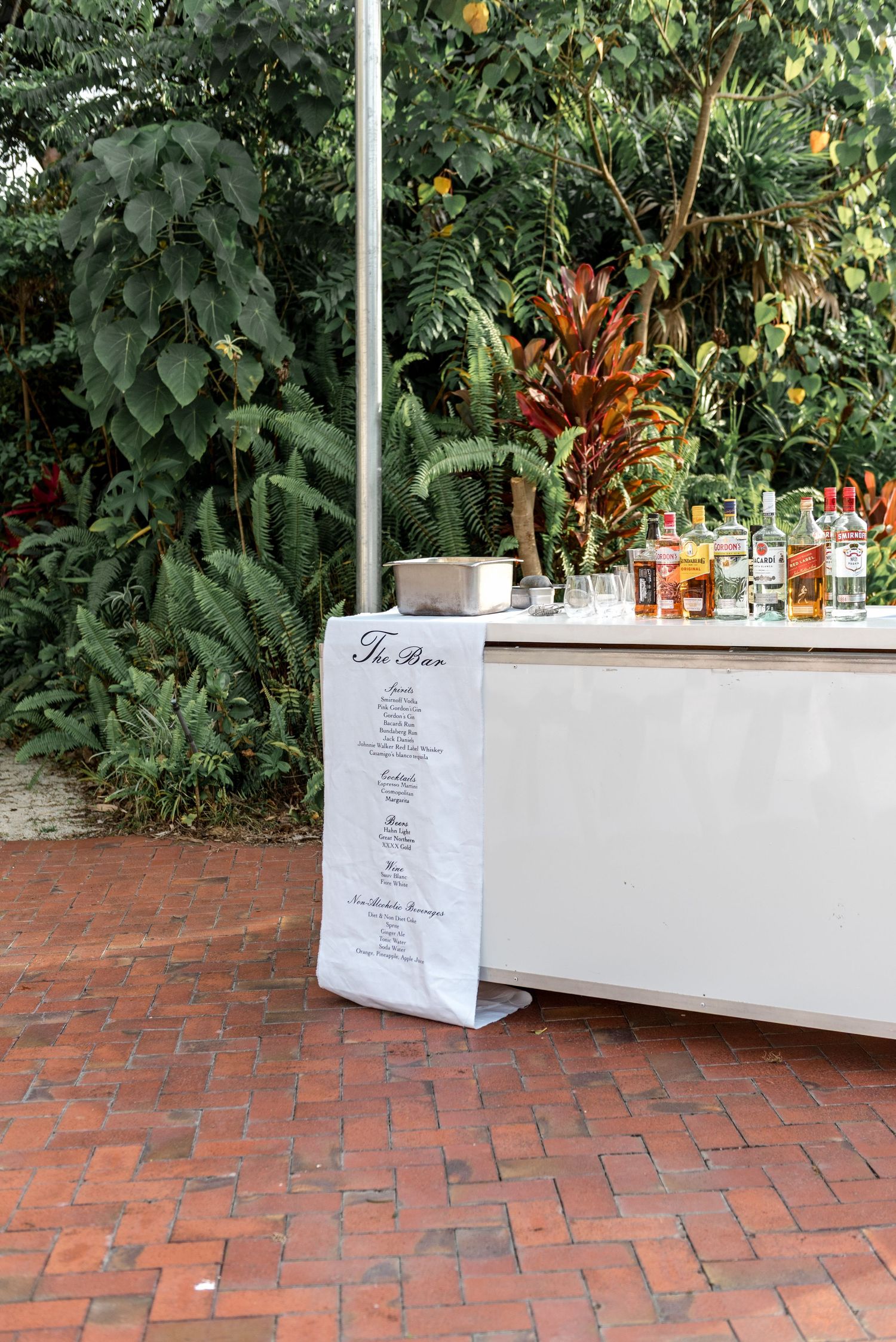 Wedding bar setup with white menu sign and bottles on brick patio with tropical foliage.