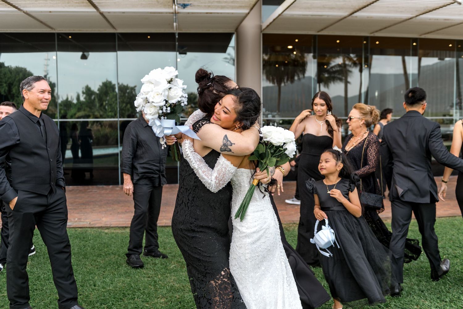Bride in white lace gown hugging guest at outdoor wedding reception with bridal party in black attire.