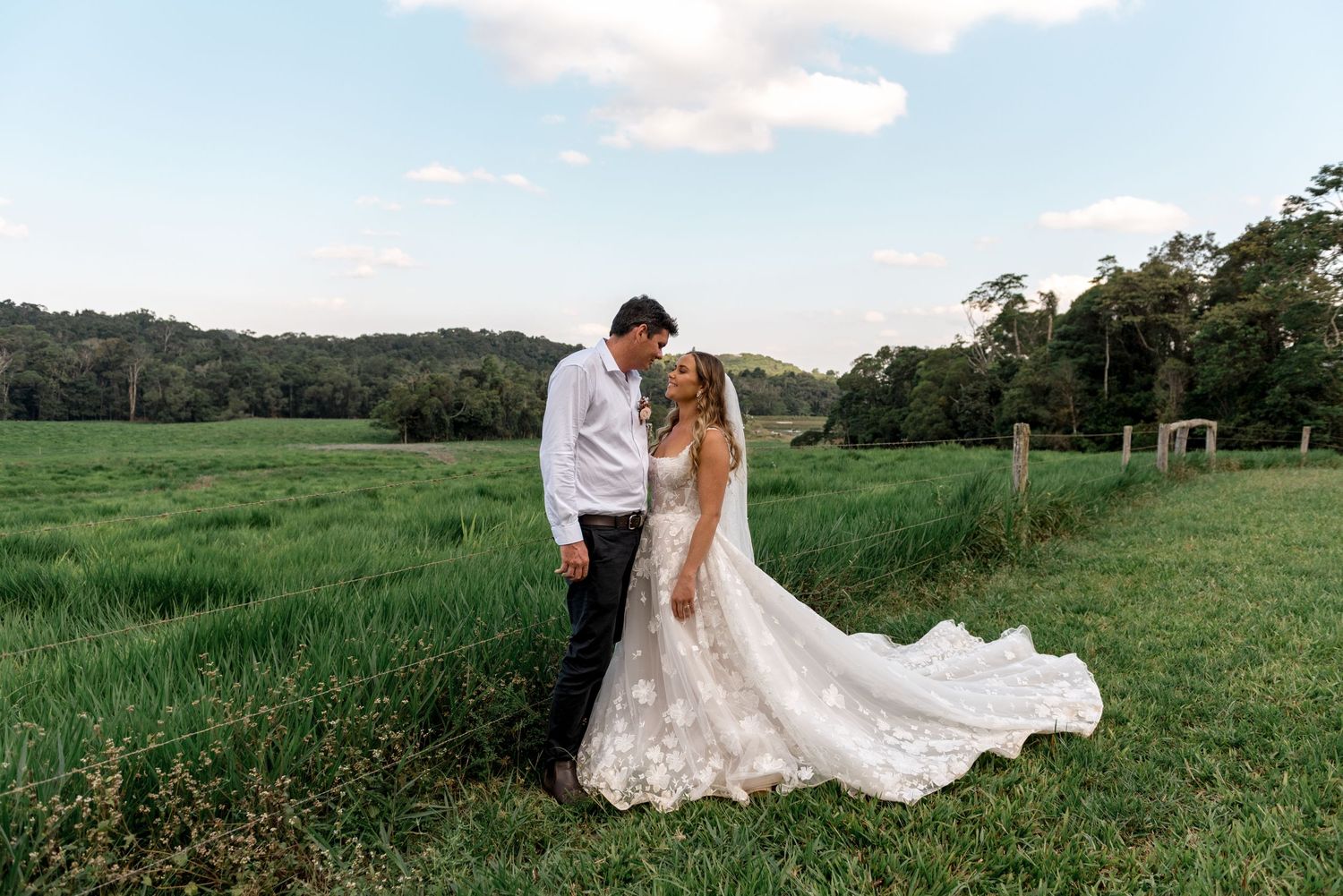 Bride and groom embracing in a lush green meadow with rolling hills and trees in background.