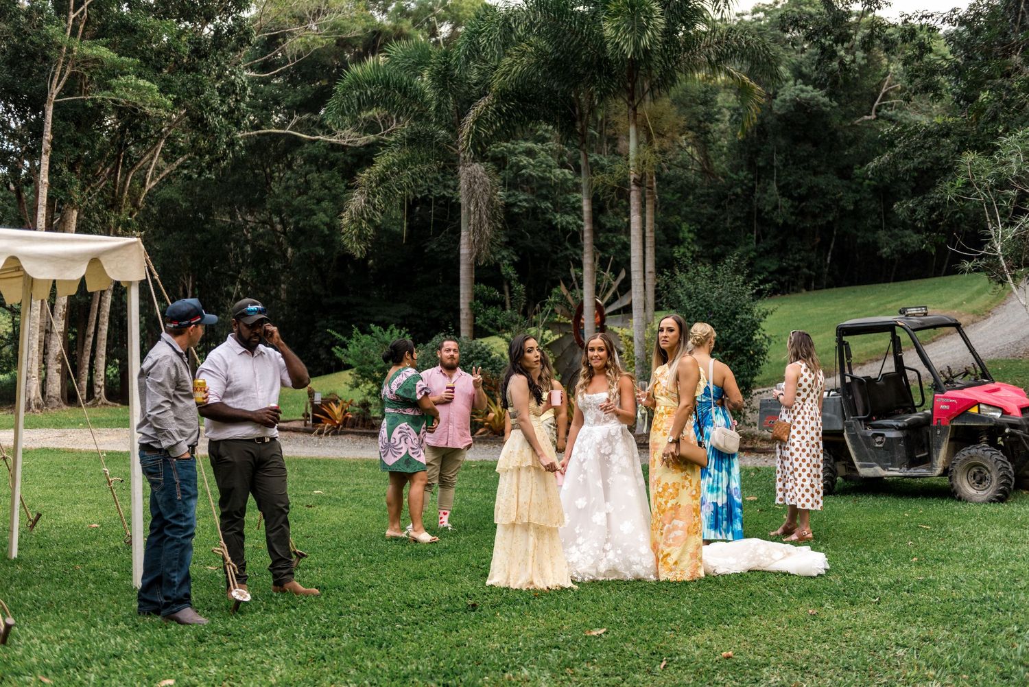 Wedding party gathering on green lawn with tropical palm trees and red utility vehicle nearby.
