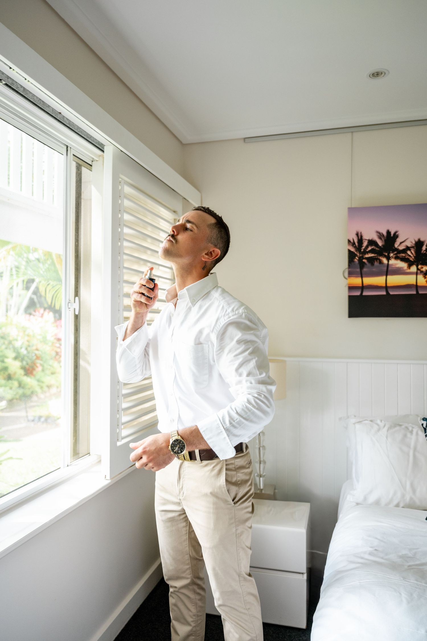 Groom in white shirt applying cologne near a sunlit bedroom window before his wedding ceremony.