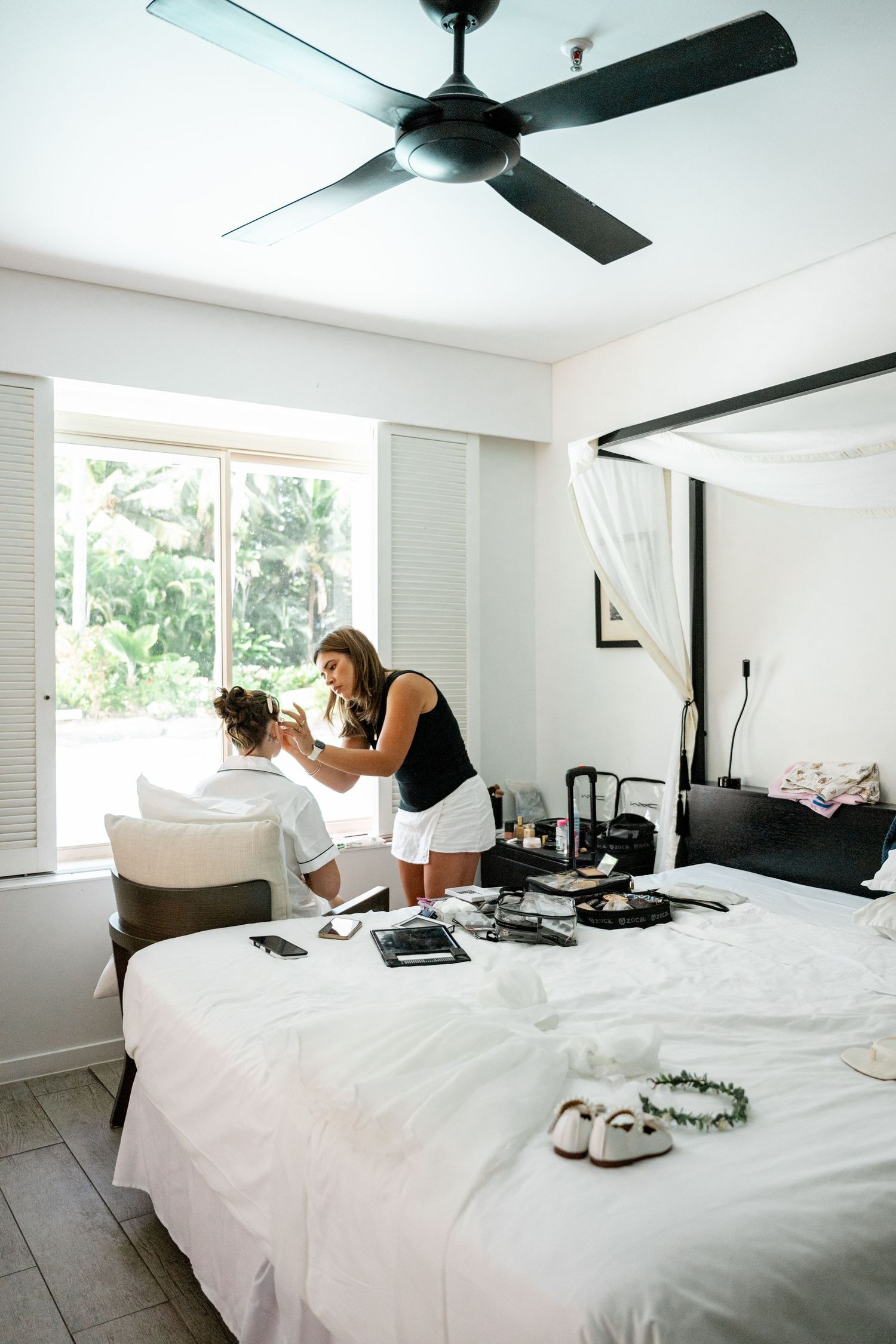 Makeup artist applying makeup to a seated woman in a bright bedroom with wedding accessories on the bed.