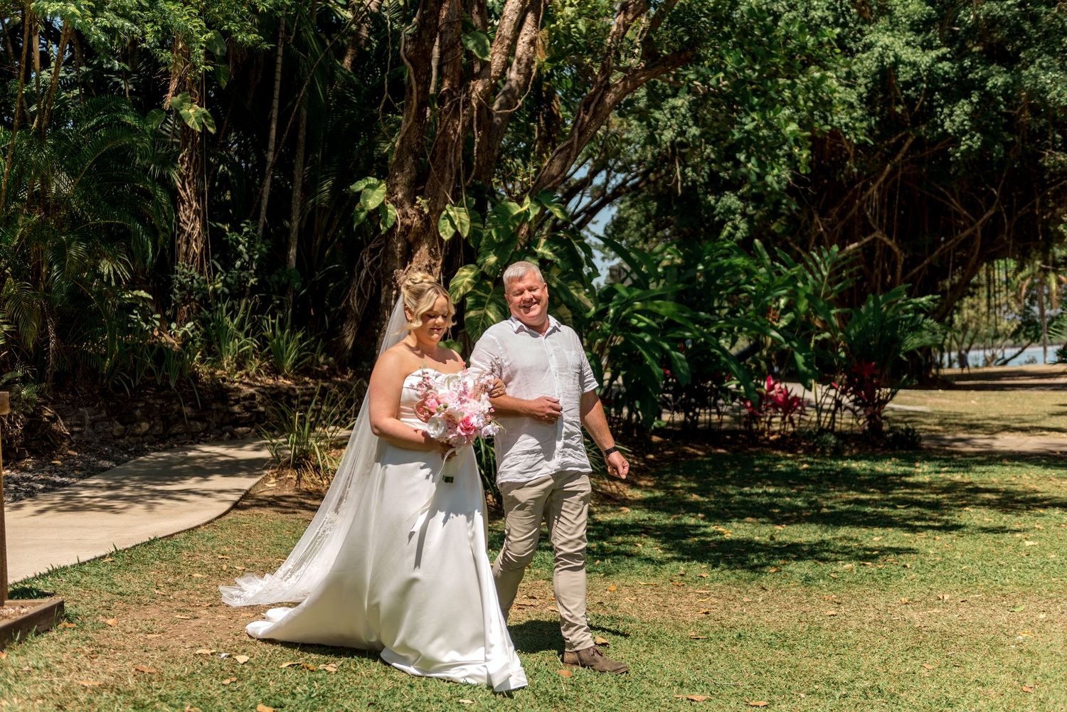 Bride in elegant white gown and groom walk through tropical garden on their wedding day.