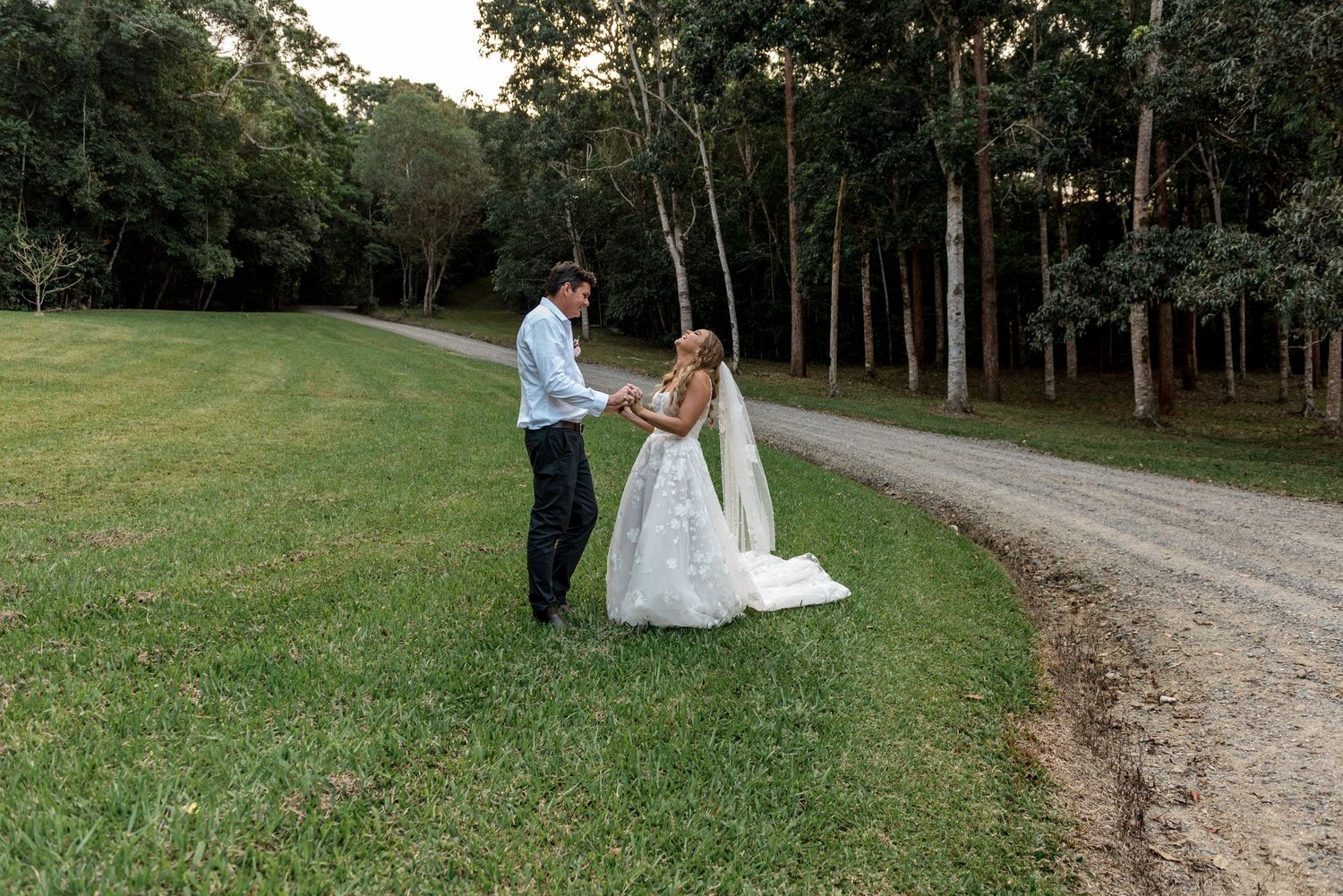Bride and groom holding hands on a green lawn next to a gravel path lined with tall trees at dusk.