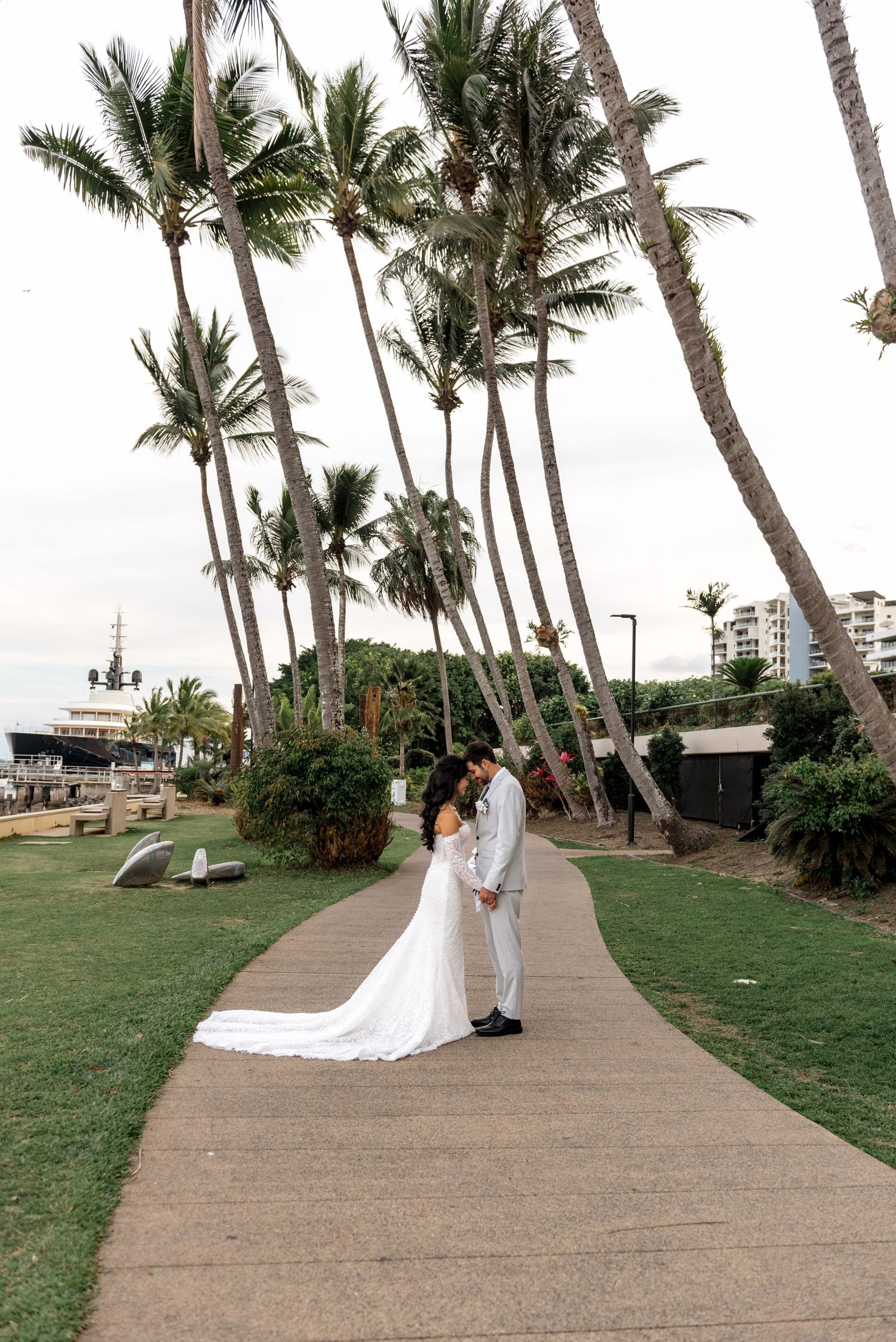 Bride and groom share intimate moment on palm-lined pathway near waterfront marina on wedding day.