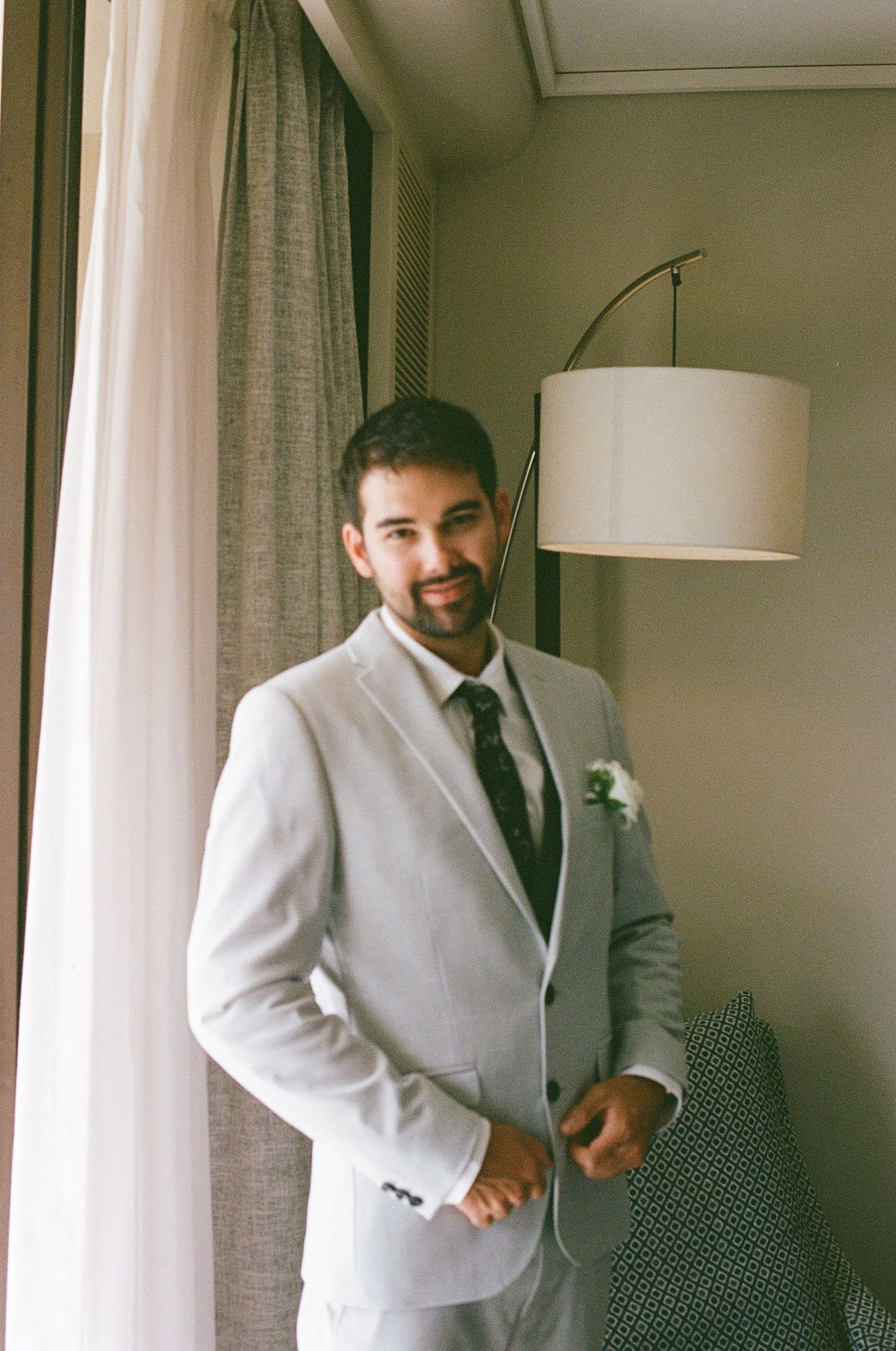Handsome groom in a light grey suit with boutonniere smiling while getting ready by a window.