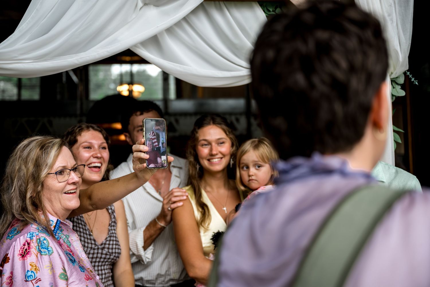 Wedding guests take a group selfie under a draped canopy at a reception.