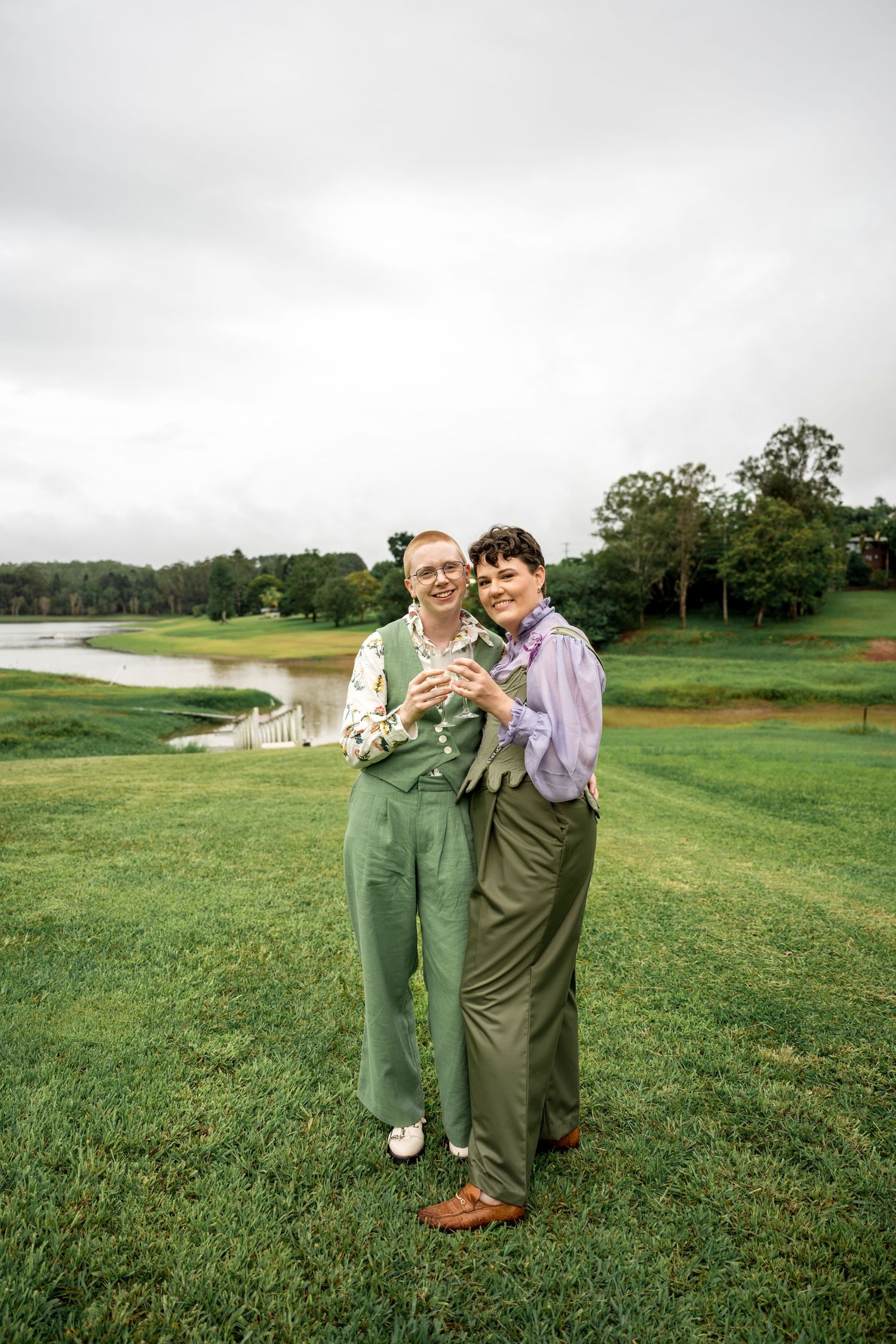 Smiling couple in coordinated green outfits embracing on a lush green lawn near a pond.