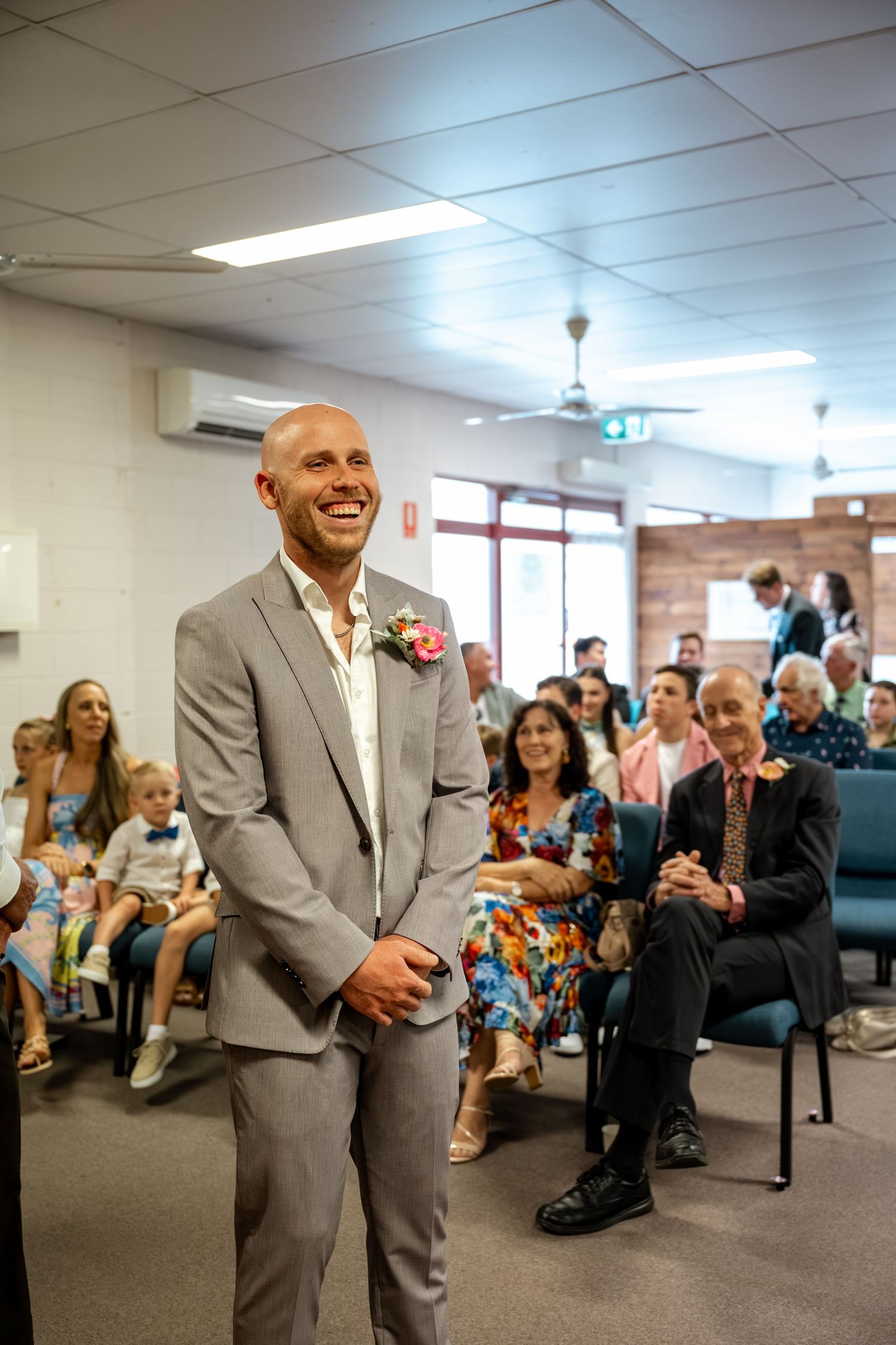Smiling groom in grey suit with floral boutonniere awaits bride at indoor wedding ceremony.