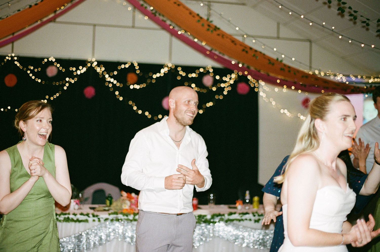 Bride and guests laughing and clapping together under colorful drapes at a lively wedding reception.