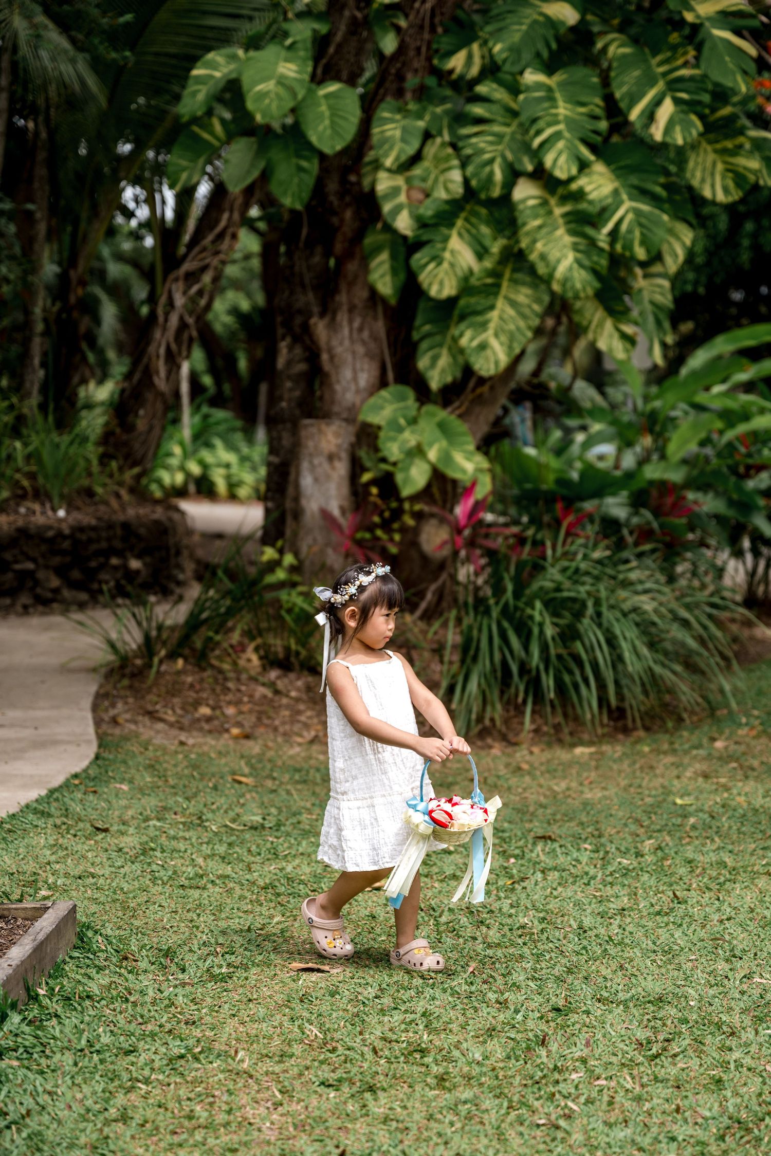 Young flower girl in white dress carries basket through lush tropical garden path at Little Cove, Port Douglas