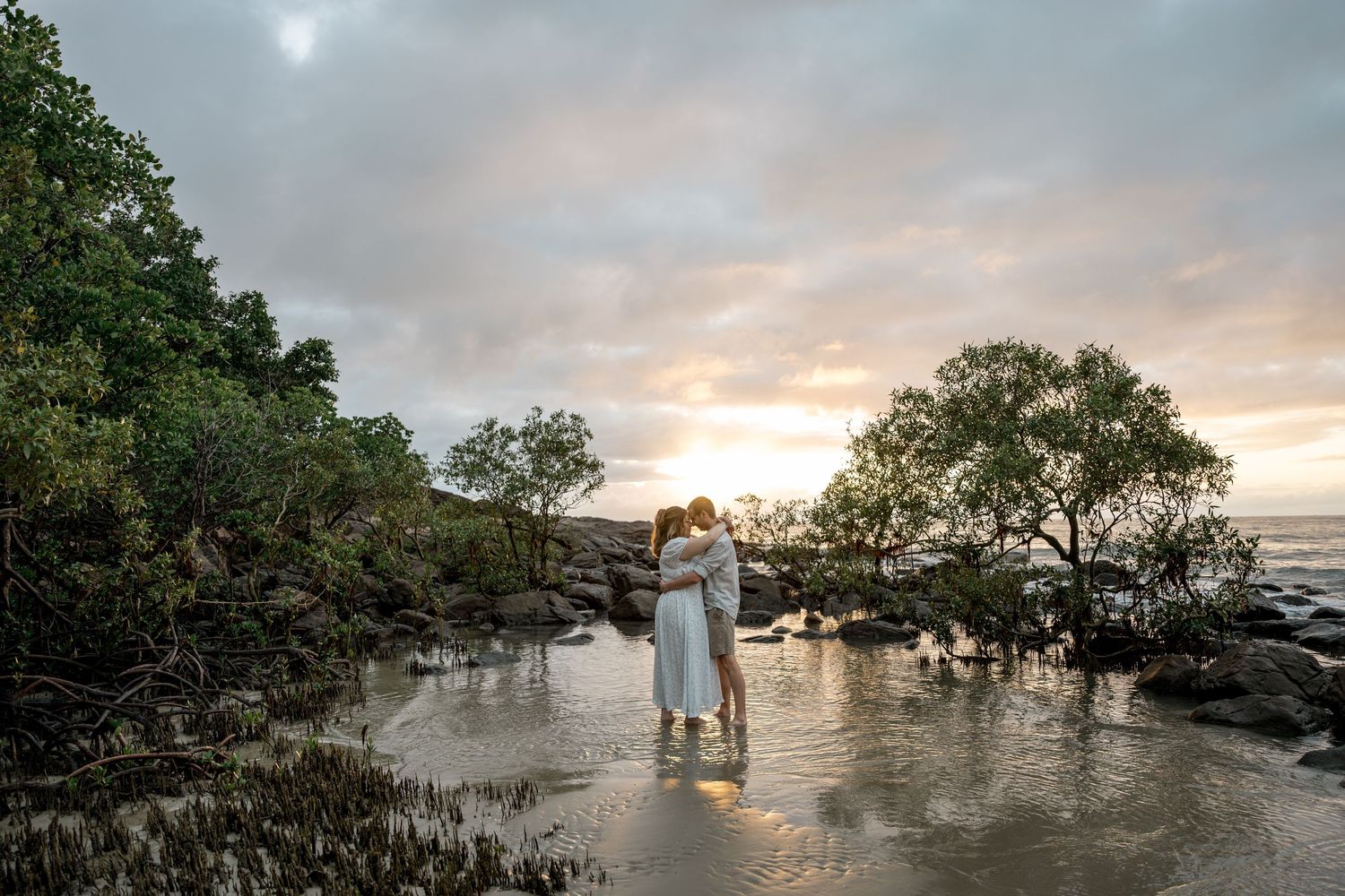 Romantic couple embracing at sunset in shallow water on a tropical beach with dramatic cloudy sky at Four Mile Beach, Port Douglas.