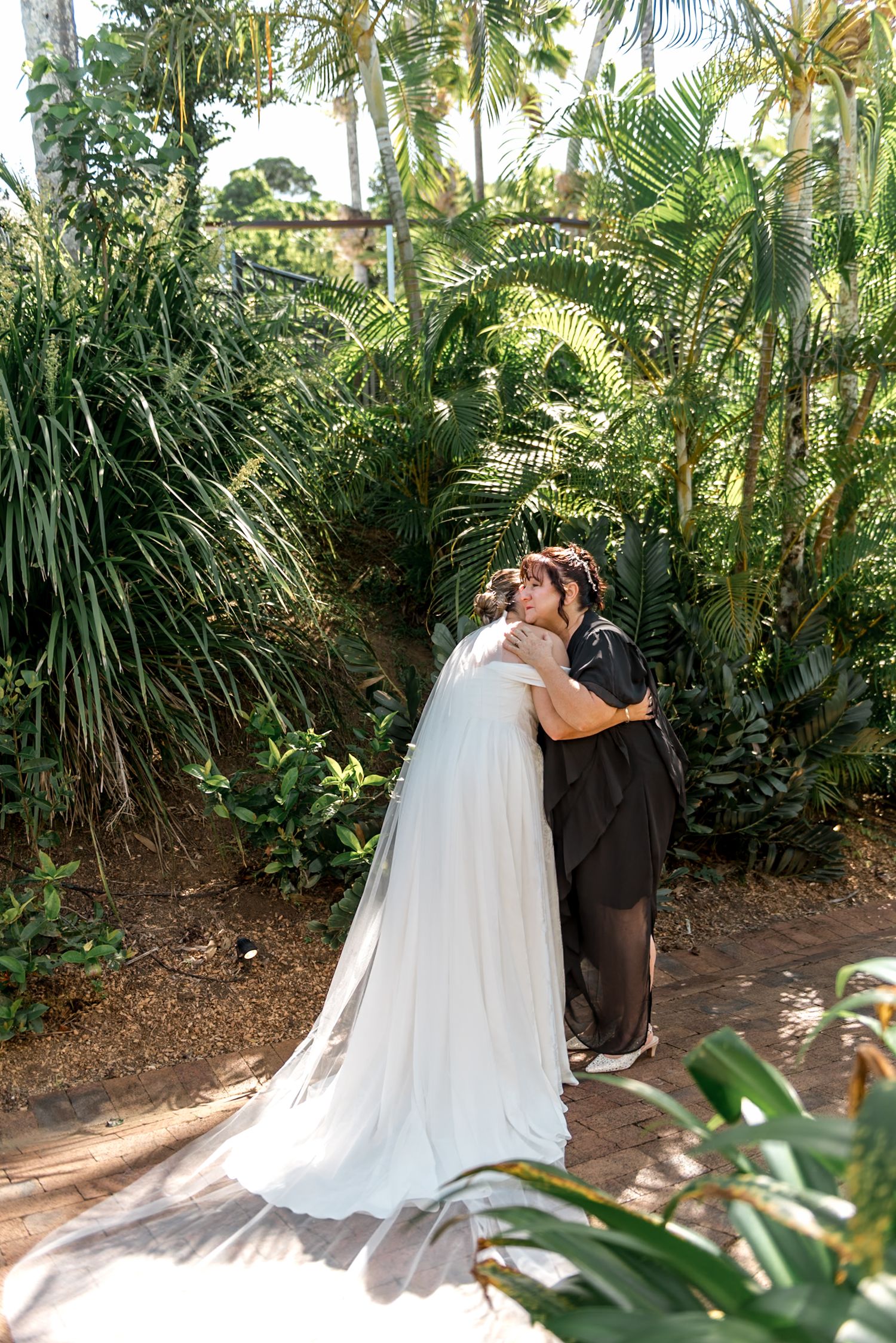Bride in white gown with long veil embraces woman in black dress in tropical garden setting.
