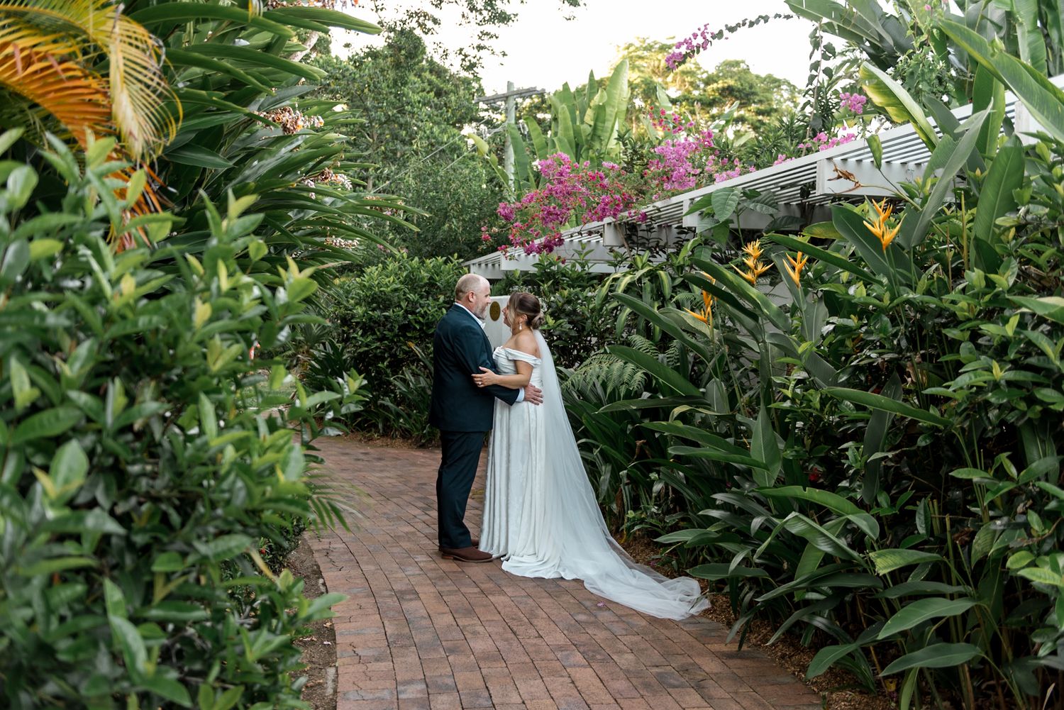 Bride and groom share intimate moment on brick pathway surrounded by lush tropical gardens.