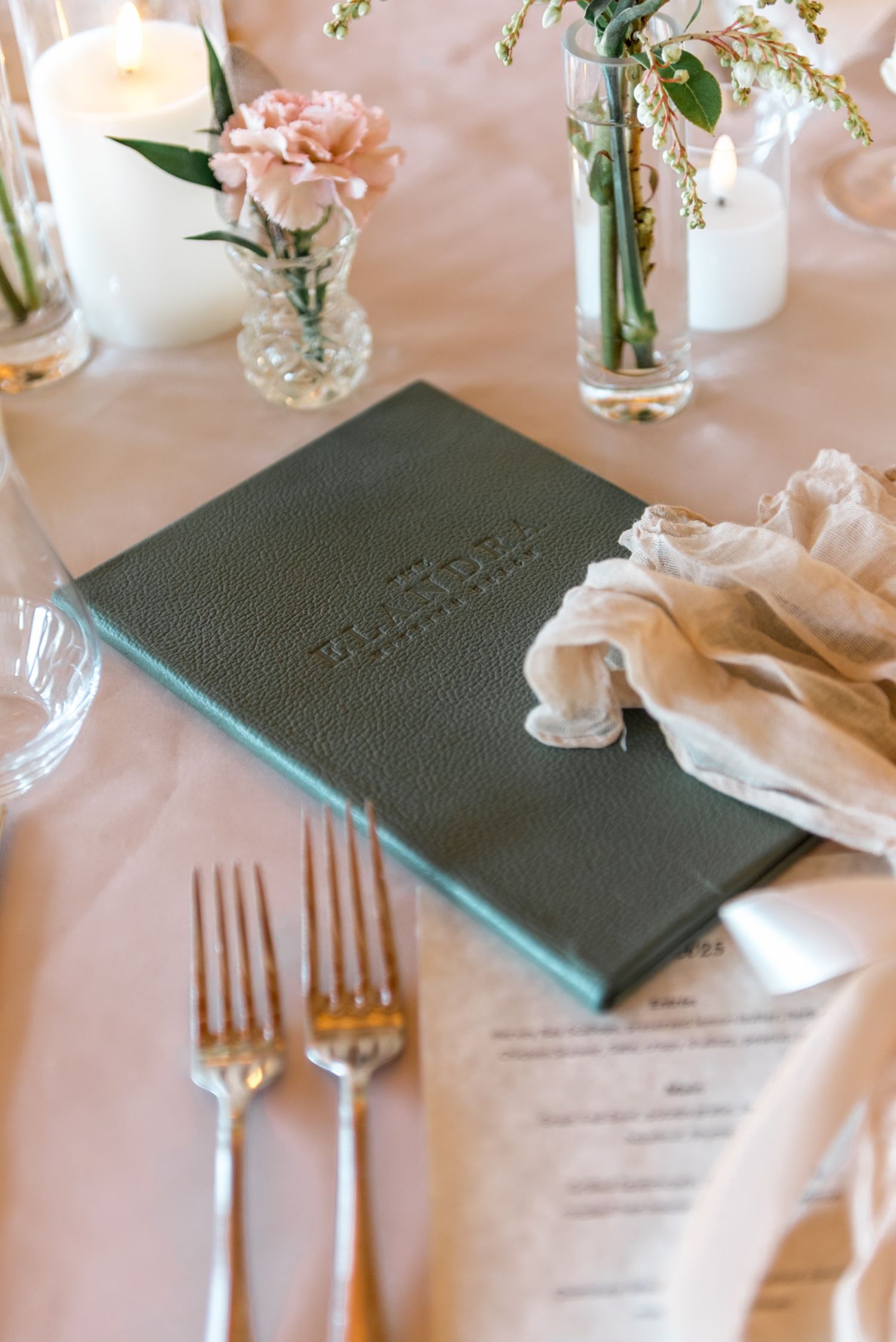 Elegant wedding place setting with green leather menu, two forks, pink carnation, and candles on blush tablecloth.