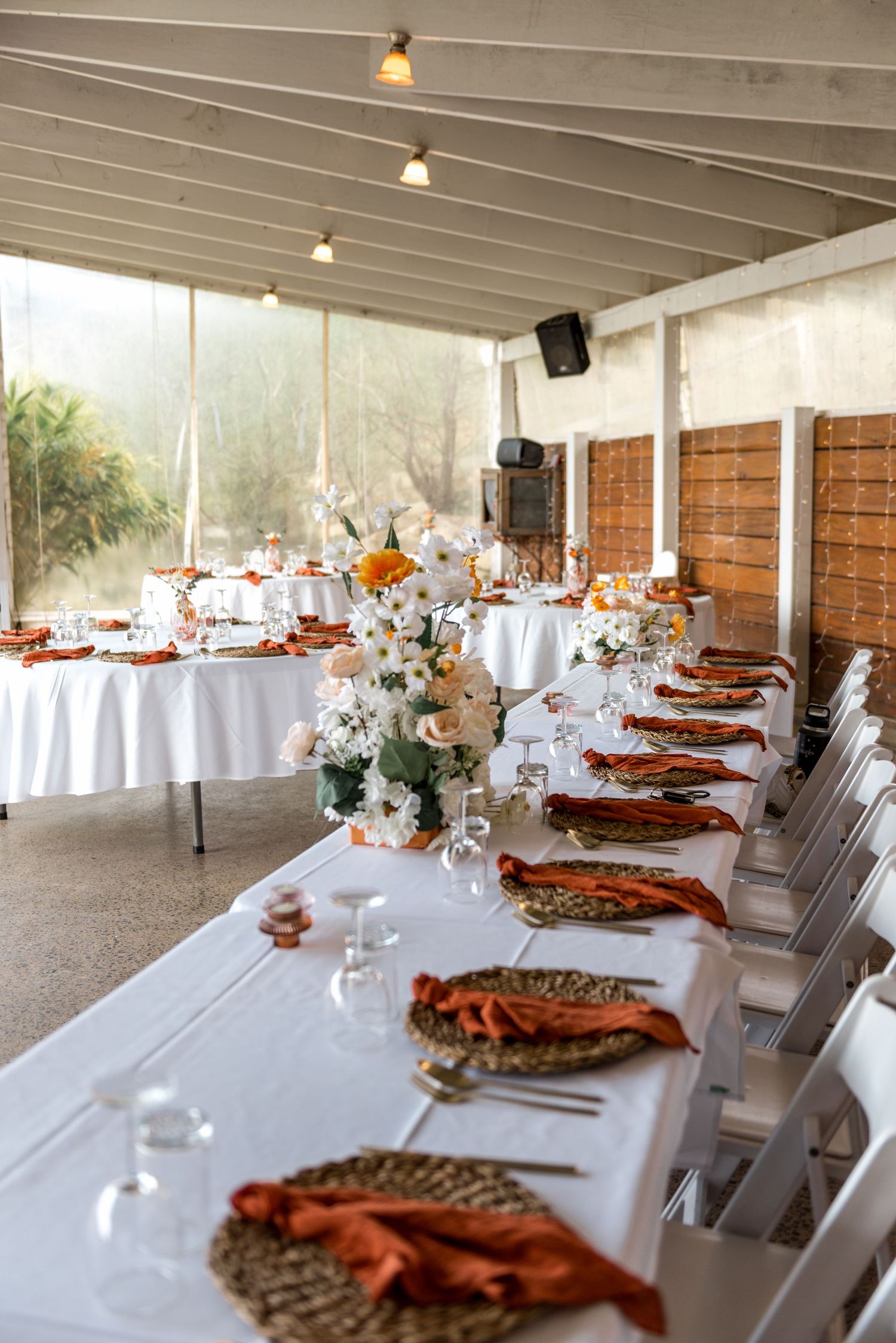 Rustic outdoor wedding reception with long white tables, woven charger plates, burnt orange napkins, and floral centerpieces.