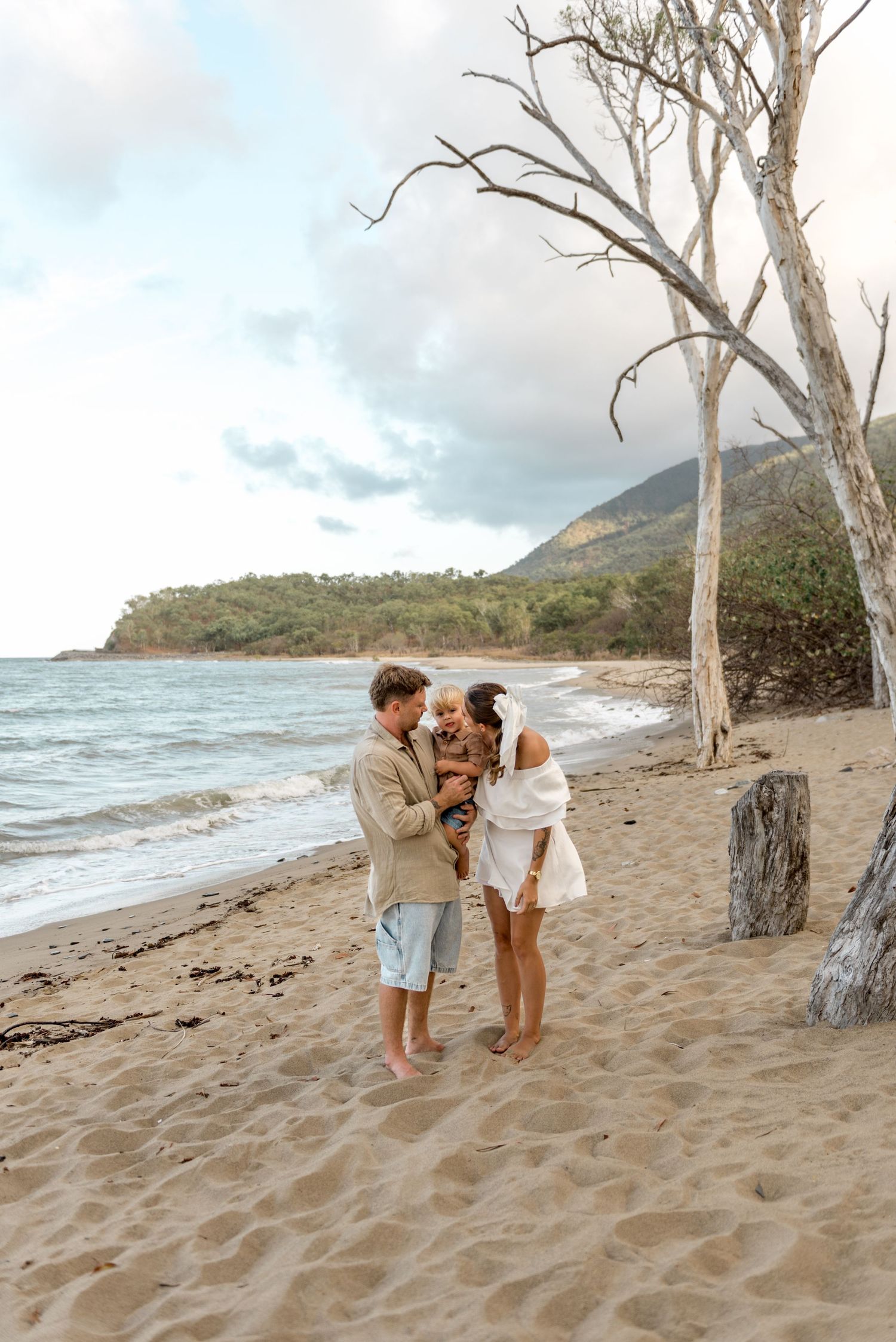 Young family laughing together on sandy beach with ocean waves and lush hillside backdrop.