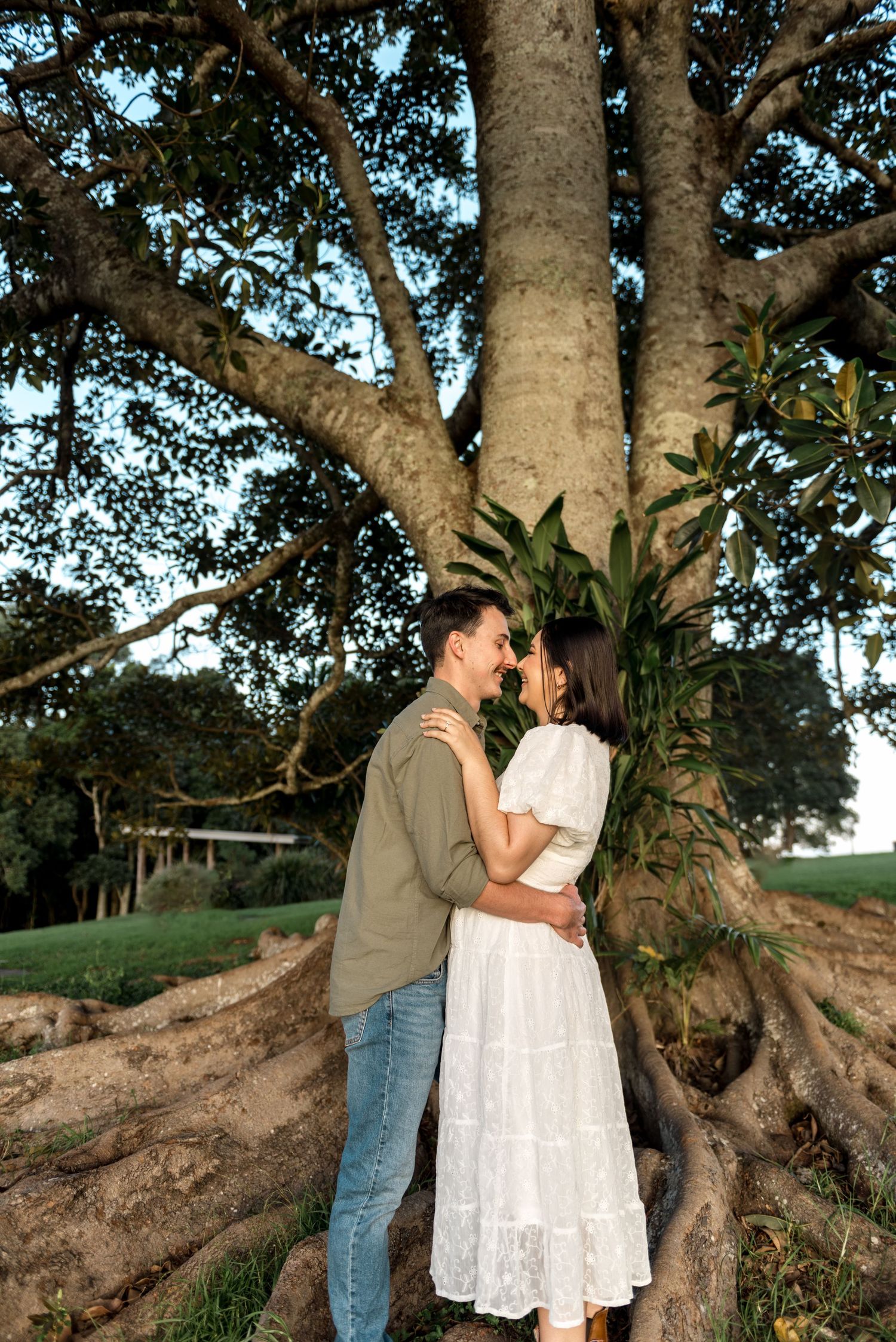 Happy couple laughing and embracing in front of large ancient tree at golden hour.