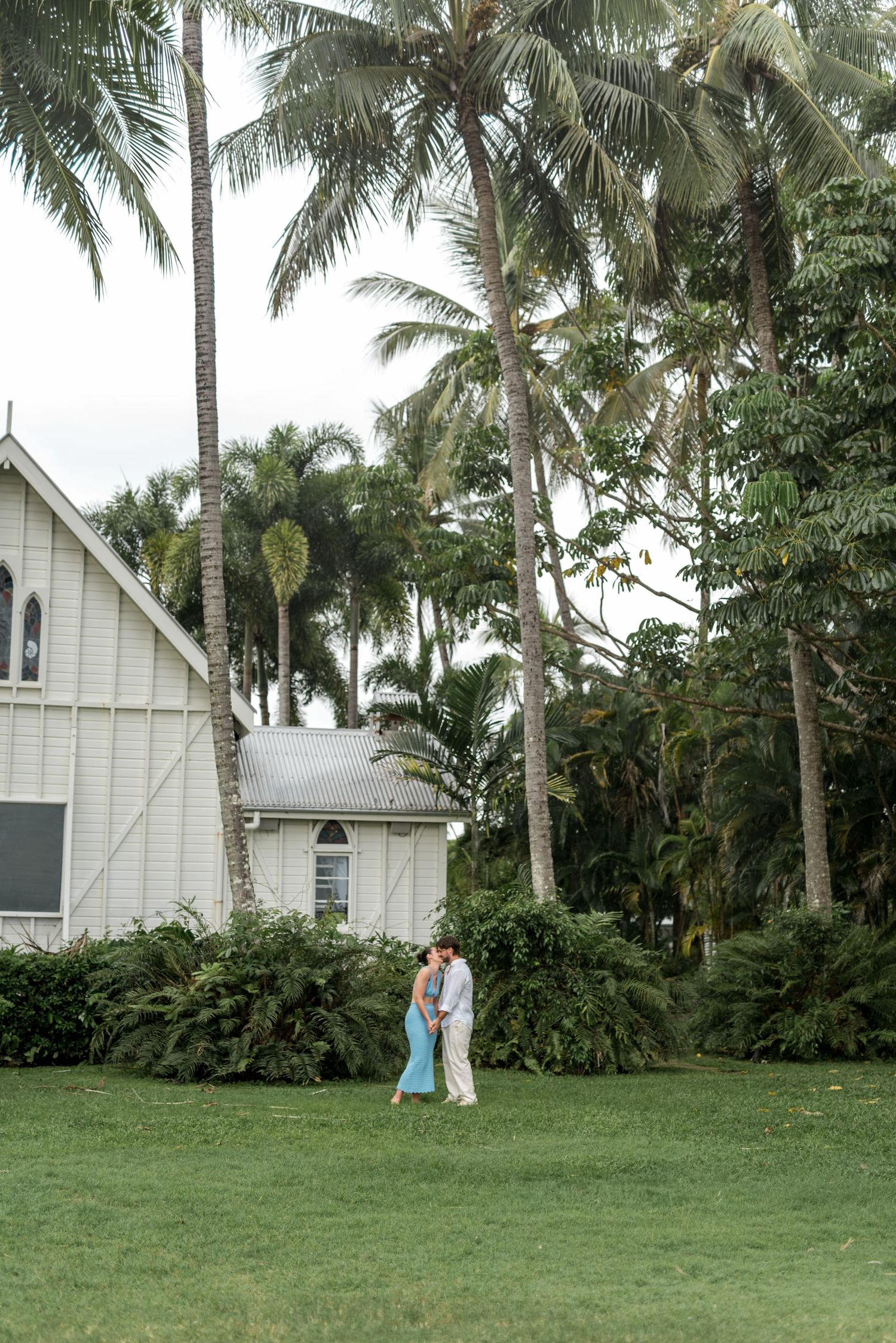 Couple kissing on green lawn surrounded by tall palm trees near a white cottage building.