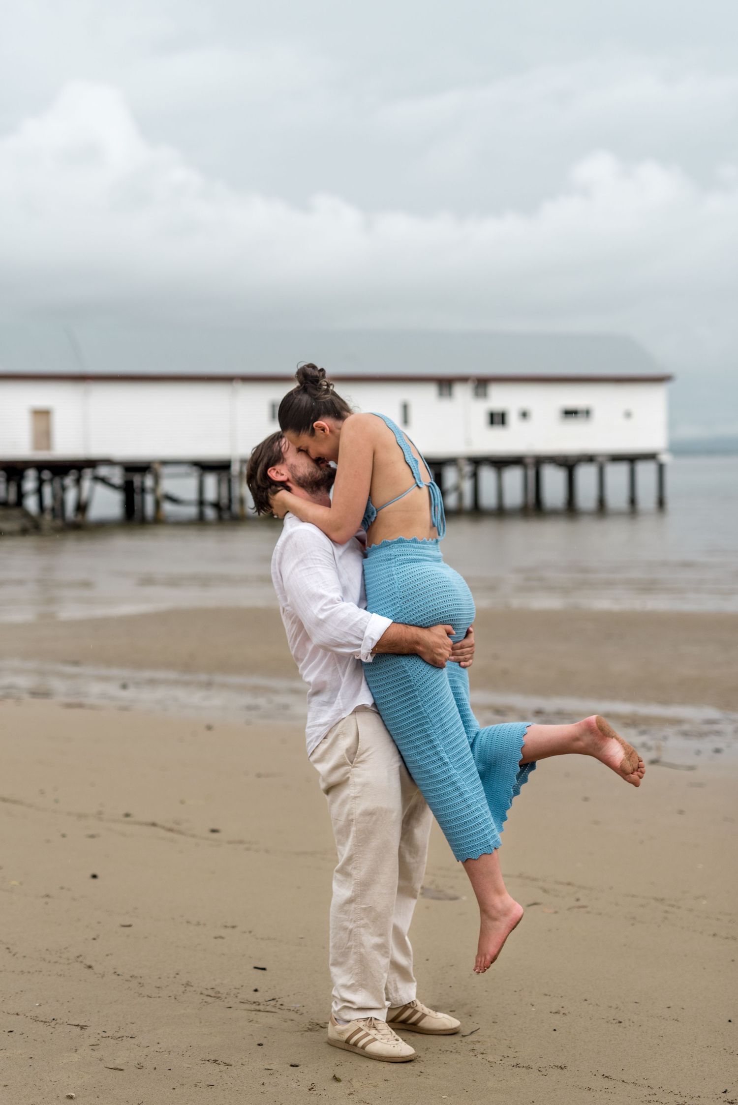 Man lifting barefoot woman in blue dress for a romantic kiss on a cloudy beach with pier.