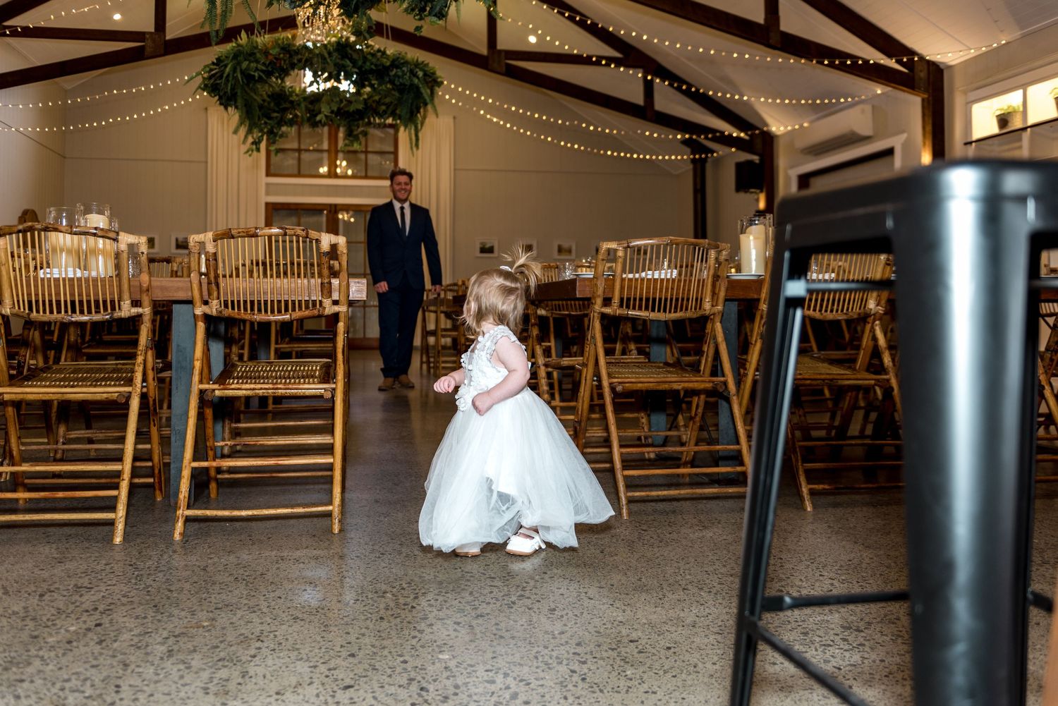Adorable flower girl in white tutu dress standing in a warmly lit rustic wedding reception barn venue.