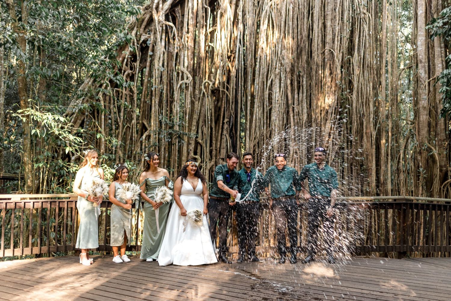 Wedding party celebrating with champagne spray in front of stunning banyan tree backdrop.