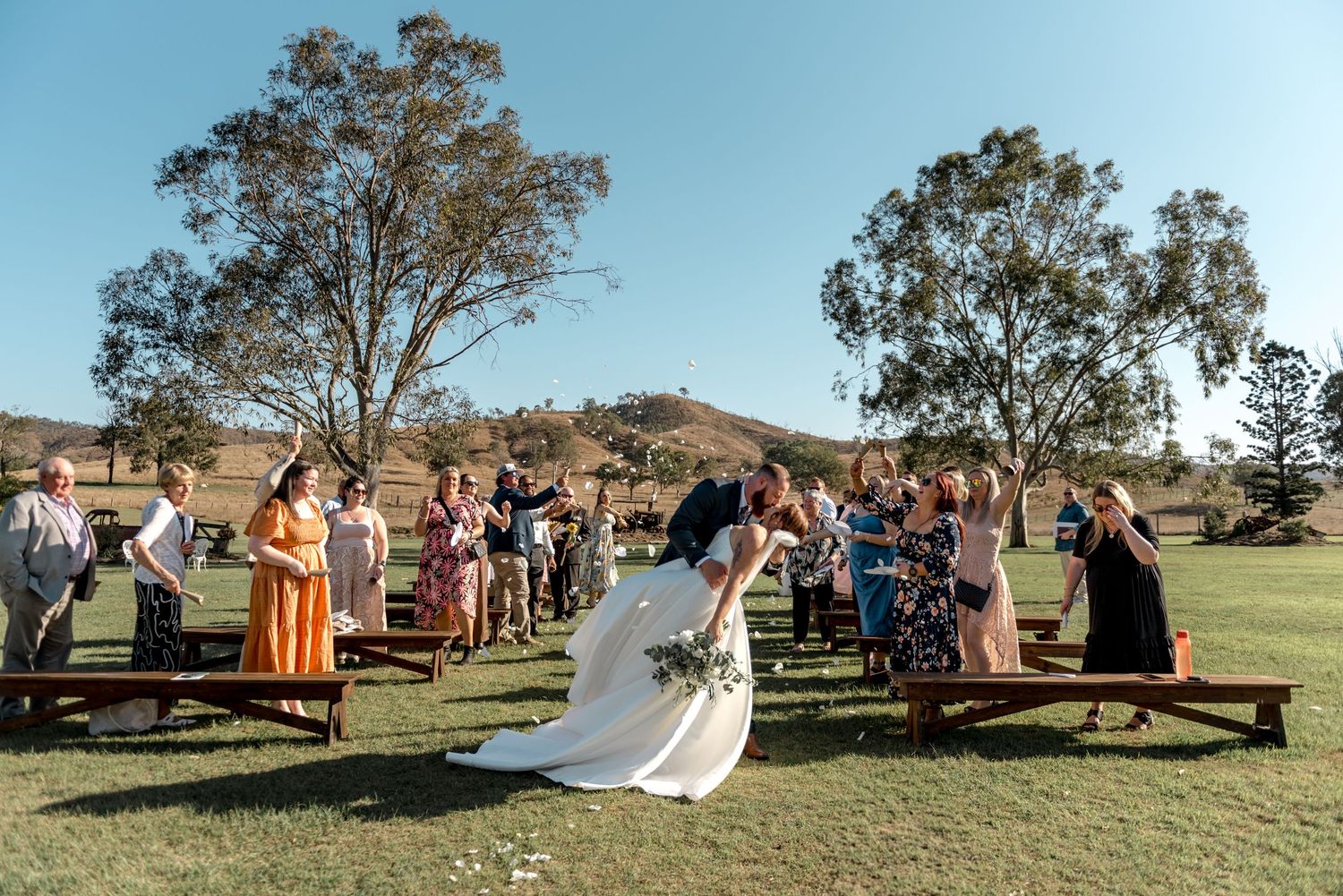 Newlyweds share a dip kiss at the end of the aisle as wedding guests celebrate at sunny outdoor ceremony.