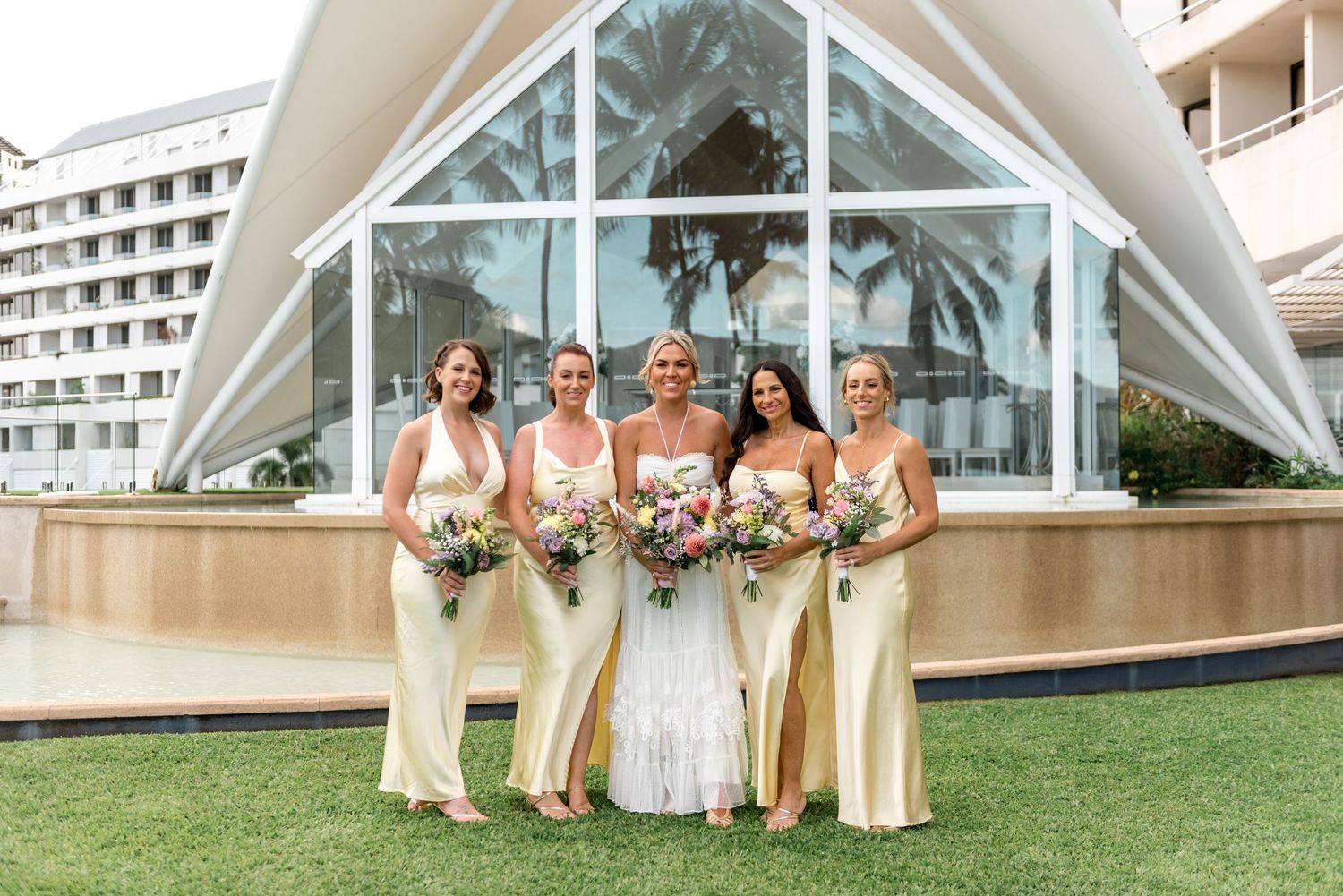 Bride and four bridesmaids in yellow dresses pose in front of a modern tropical wedding chapel.