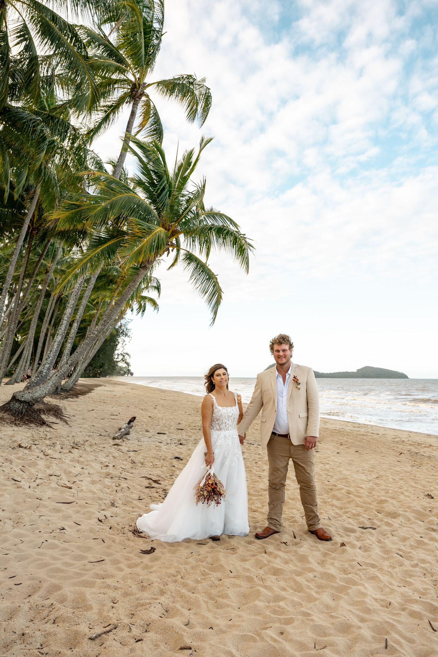 Bride and groom holding hands on a tropical beach with palm trees and ocean views on their wedding day.