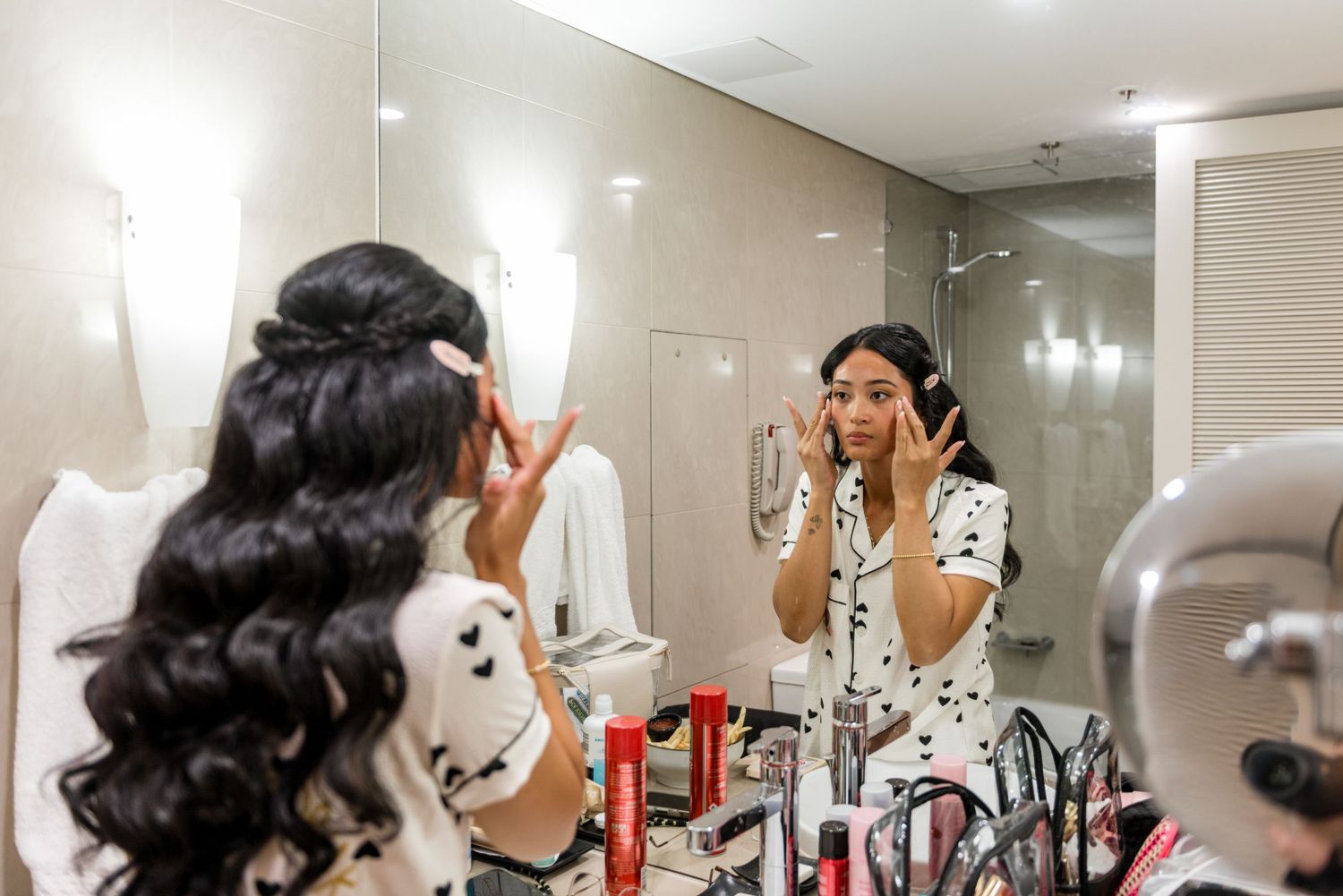 Bride applying makeup at a vanity mirror surrounded by cosmetics, wearing a black and white robe before wedding.