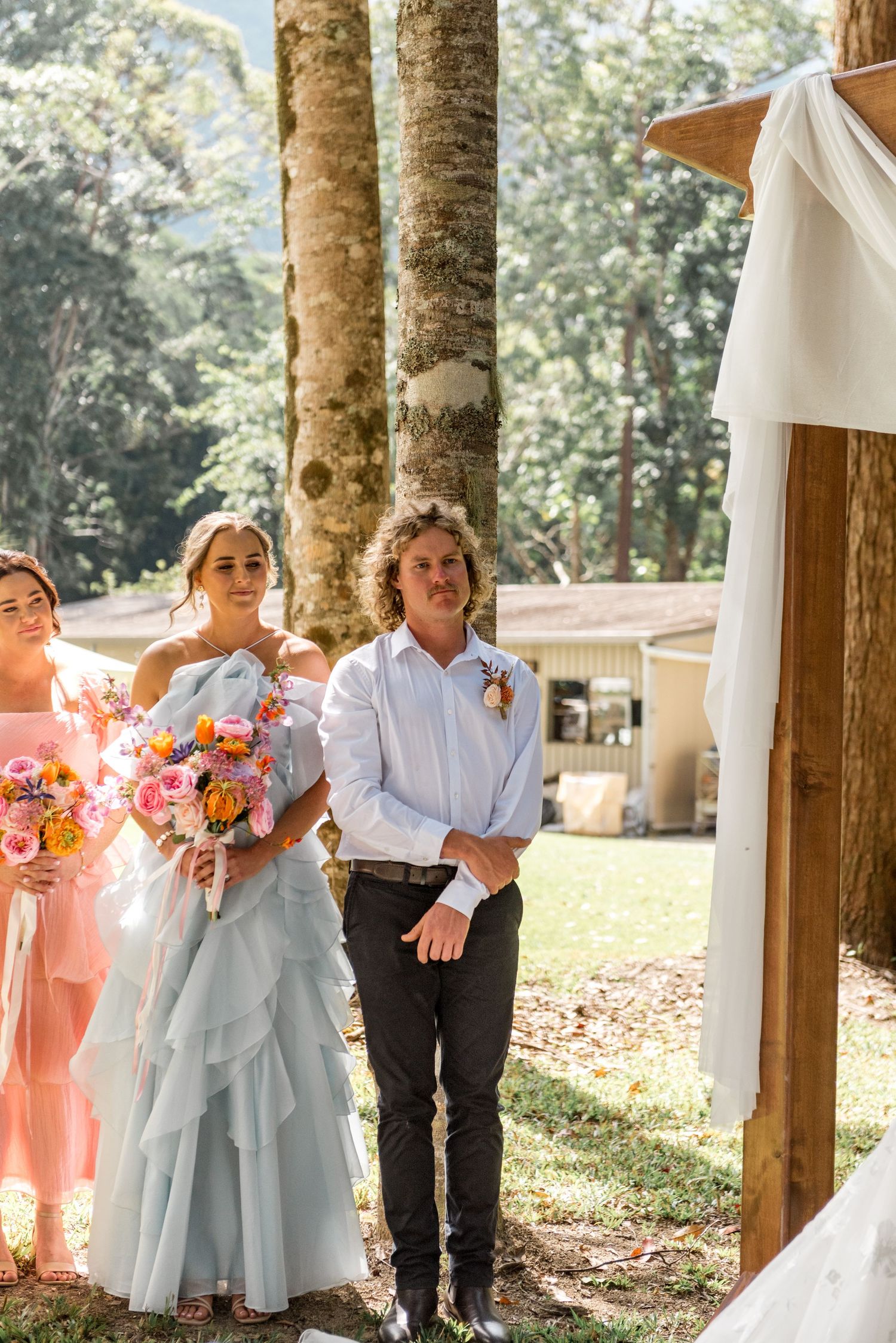 Groom watches bride approach wooden arch at outdoor forest wedding ceremony with bridesmaids.