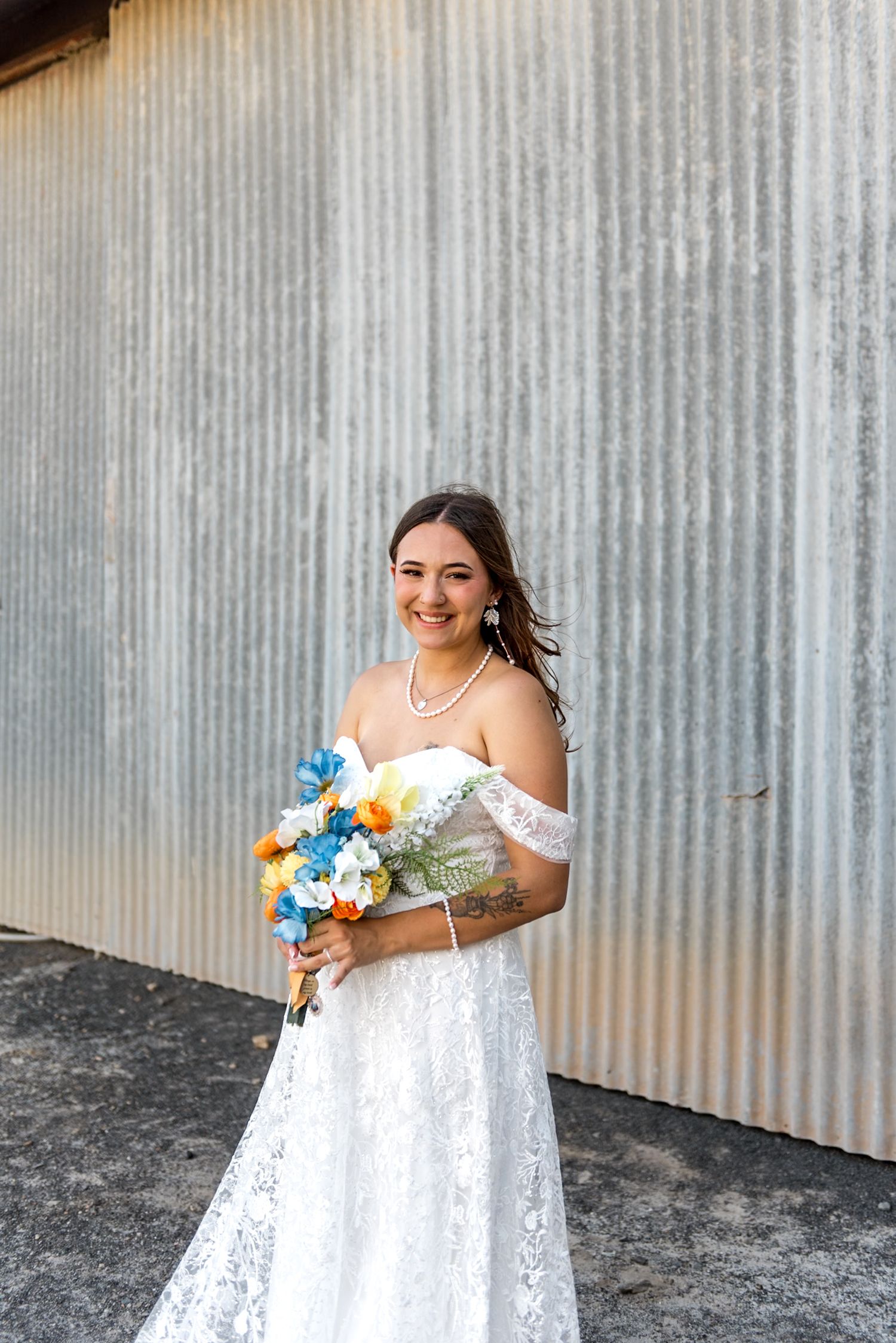 Happy bride in off-shoulder lace wedding dress holds vibrant blue and orange bouquet against metal wall.
