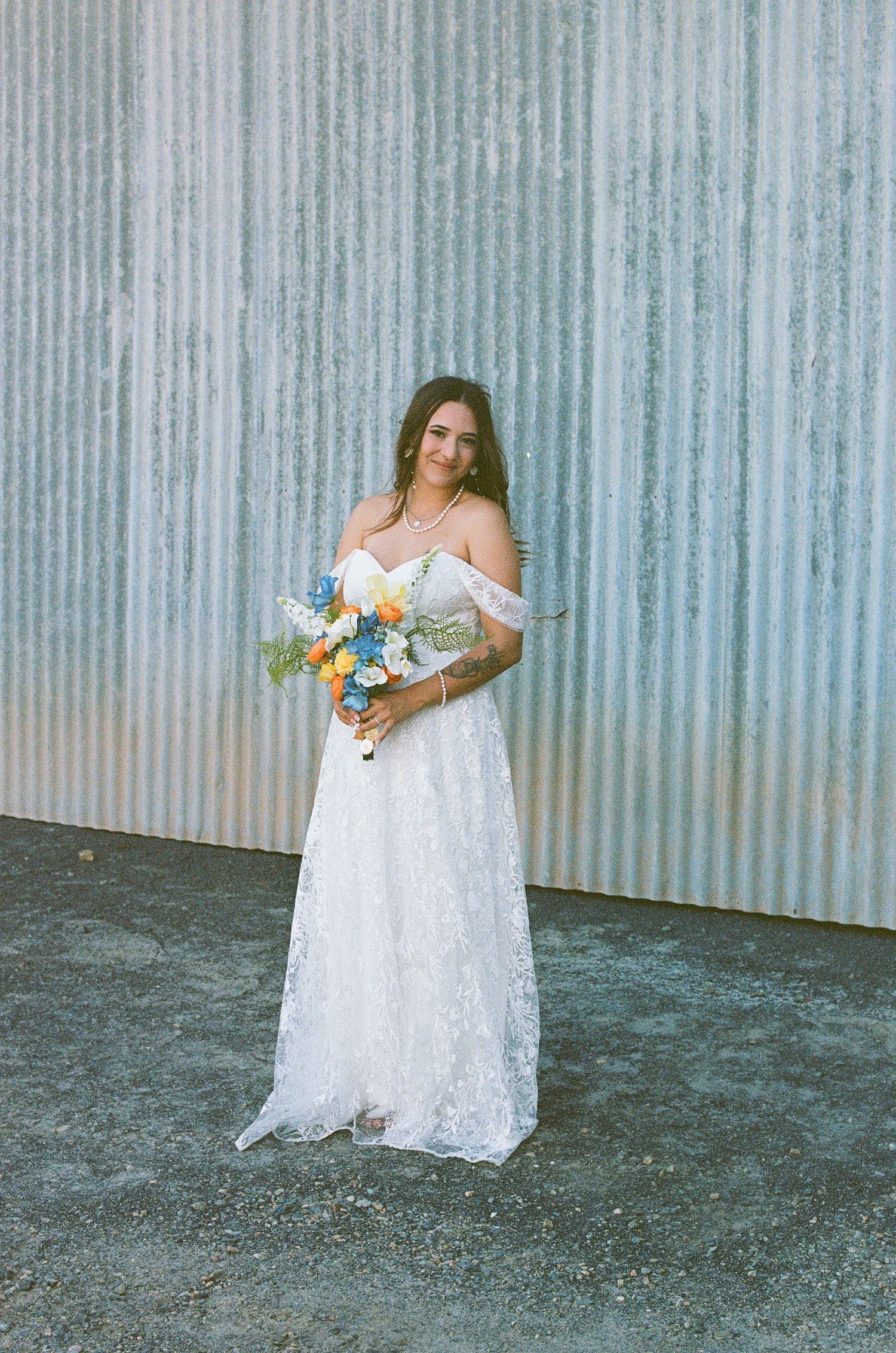 A smiling bride in an off-shoulder lace gown holds a vibrant blue and orange bouquet against a corrugated metal wall.