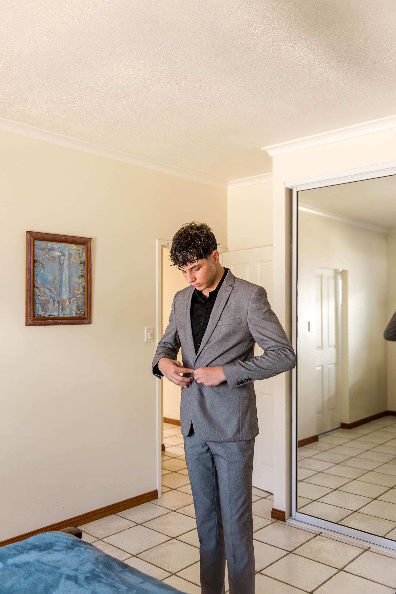 Young groomsman in grey suit buttoning jacket while getting ready in bedroom.