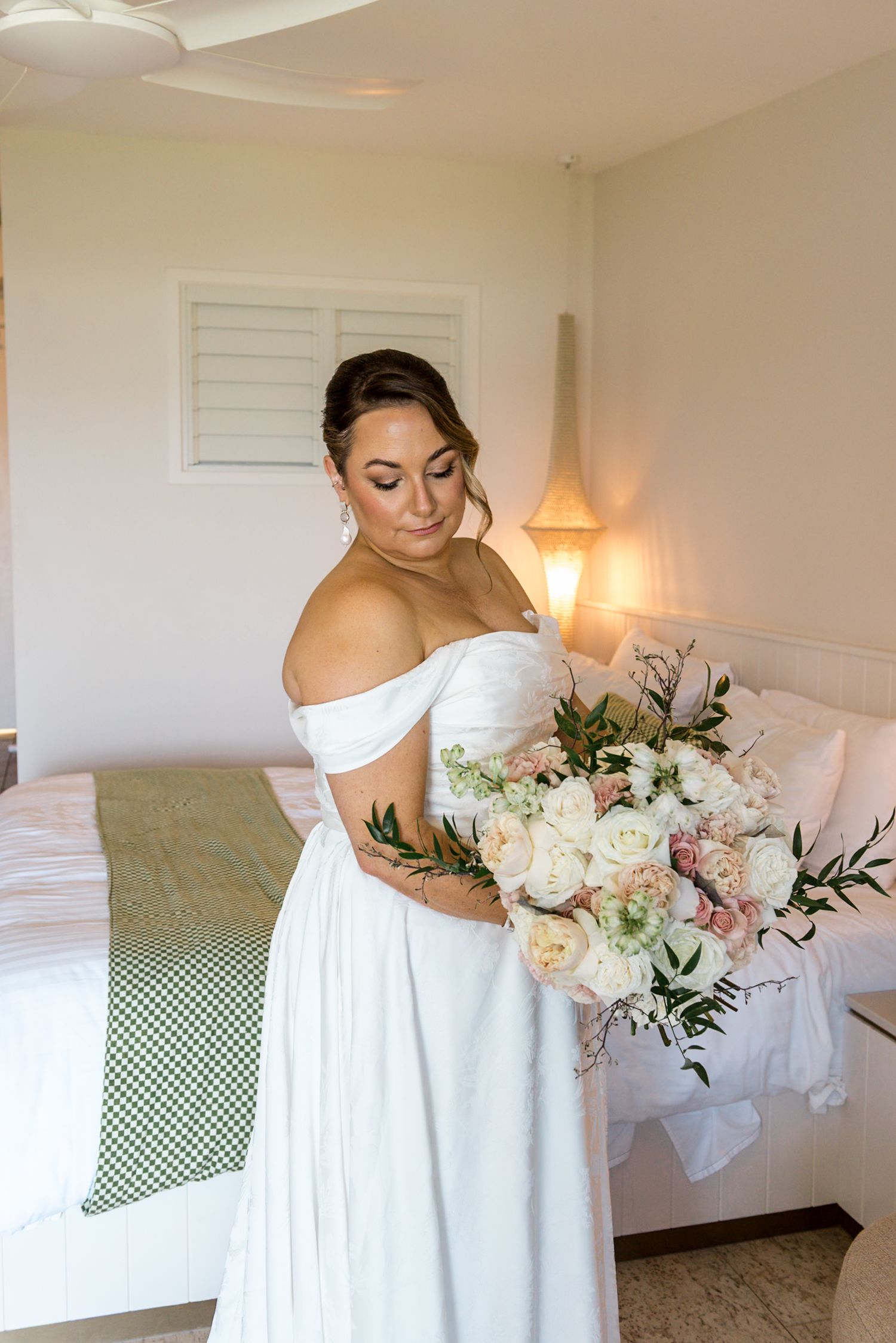Bride in off-shoulder white gown holding a lush bouquet of white and blush roses in a hotel room.