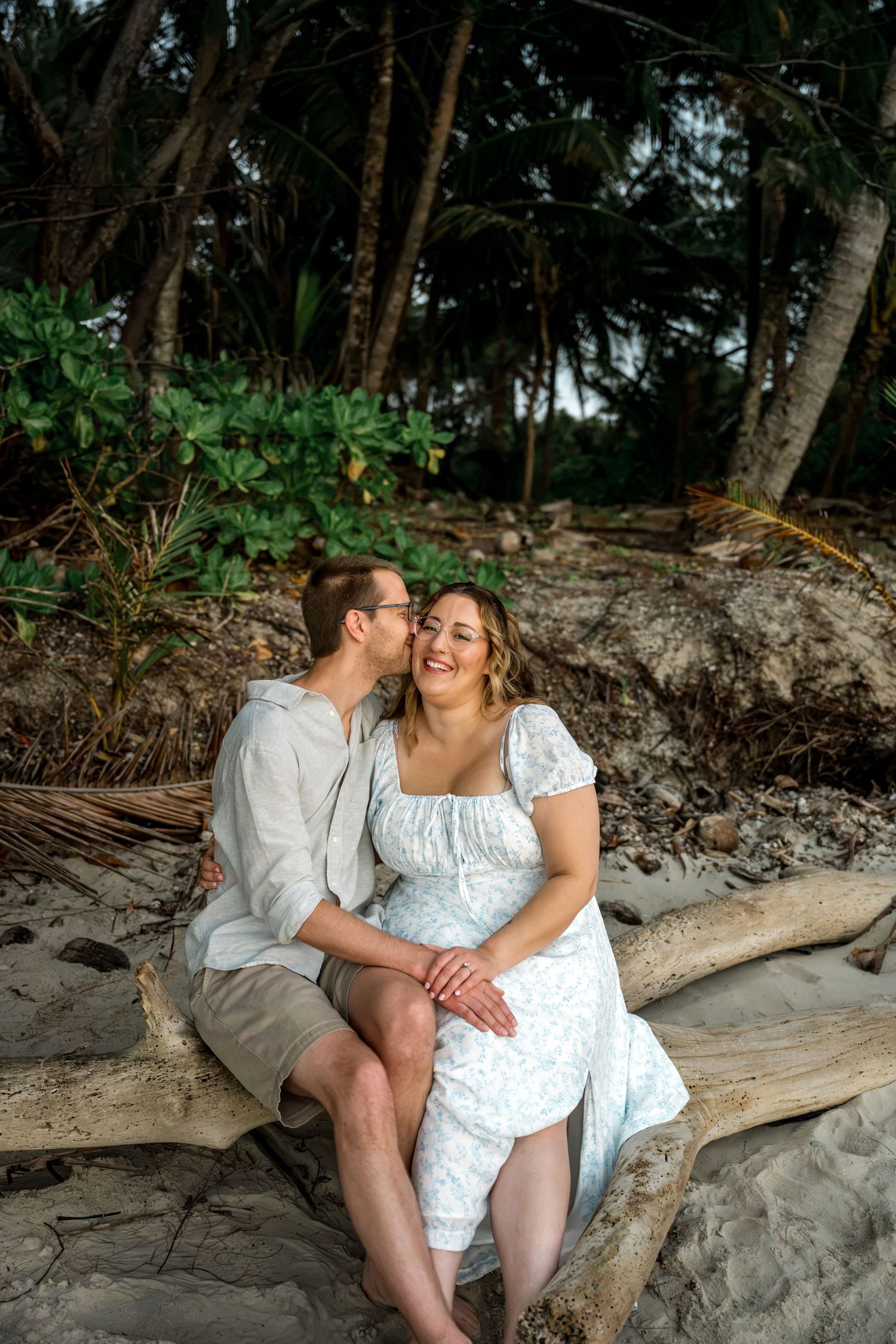 Man kissing woman on cheek as she laughs joyfully while sitting on driftwood on a tropical beach at Four Mile Beach, Port Douglas.