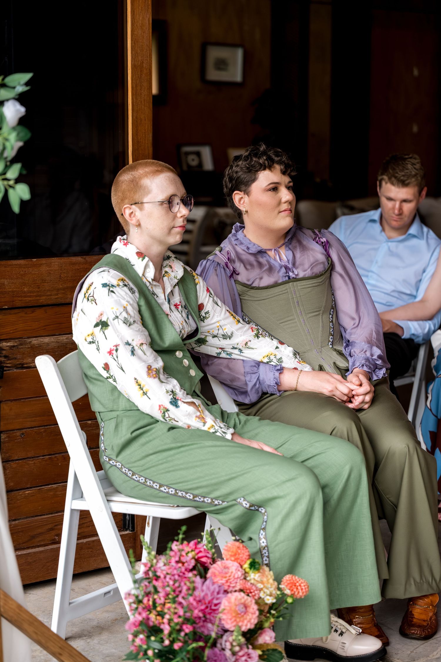 Two brides in green outfits sitting together holding hands during wedding ceremony on white chairs.