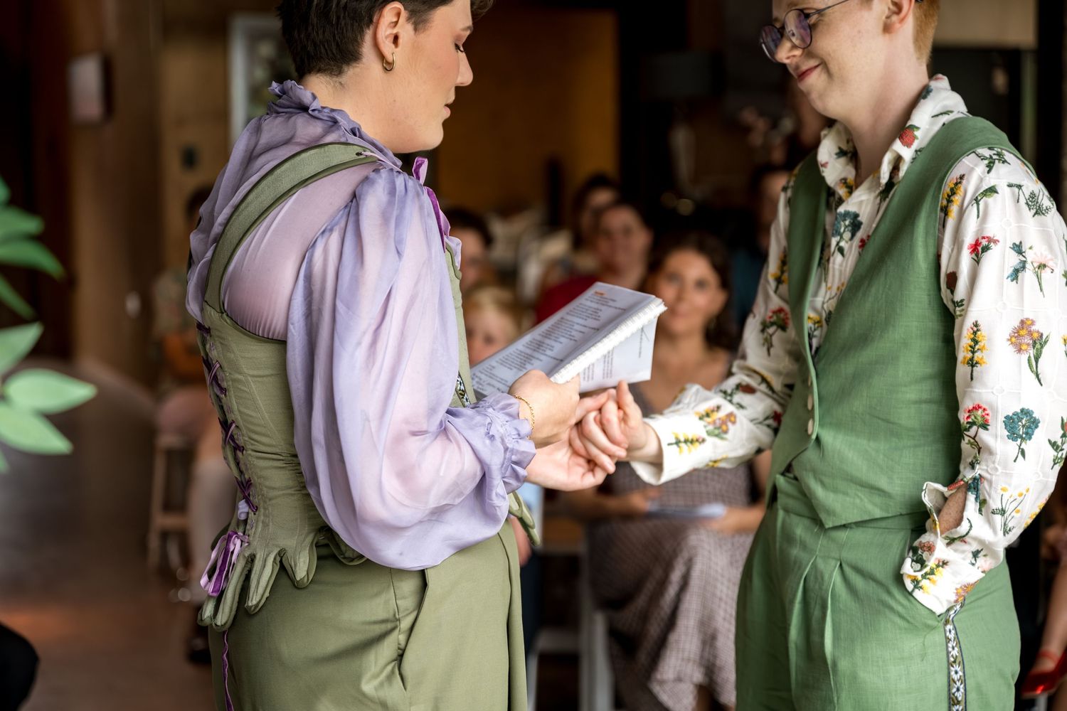 Two brides exchange rings during their wedding ceremony in coordinating green outfits.
